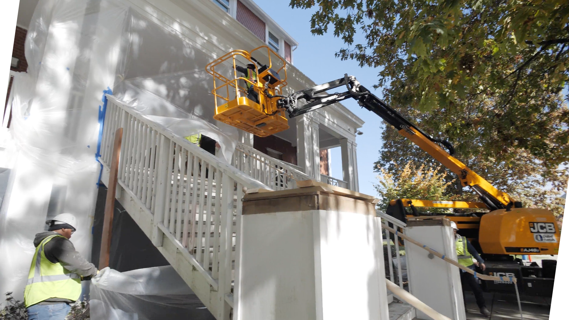 Construction workers wearing safety vests and helmets operate a cherry picker lift to perform work on the exterior of a residential house. The house has white siding and a porch with a white railing. Workers are visible on the lift working near the house's wall, with one worker on the ground assisting.