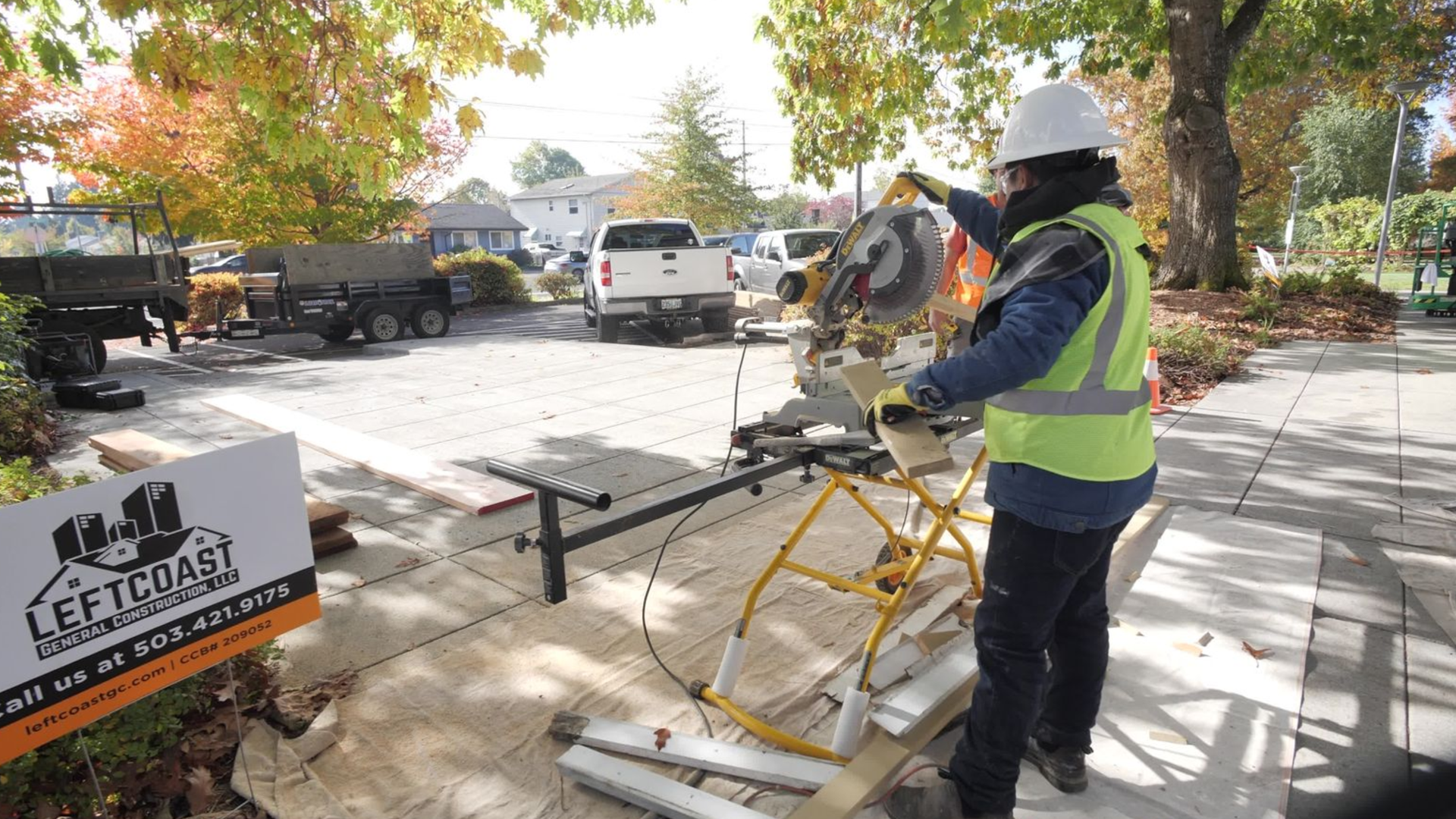 Construction worker with a hard hat and high-visibility vest cutting a piece of wood on a concrete sidewalk with a chop saw. There are trees with autumn foliage, parked cars, and construction equipment in the background. A sign for Left Coast General Construction LLC is visible in the foreground.