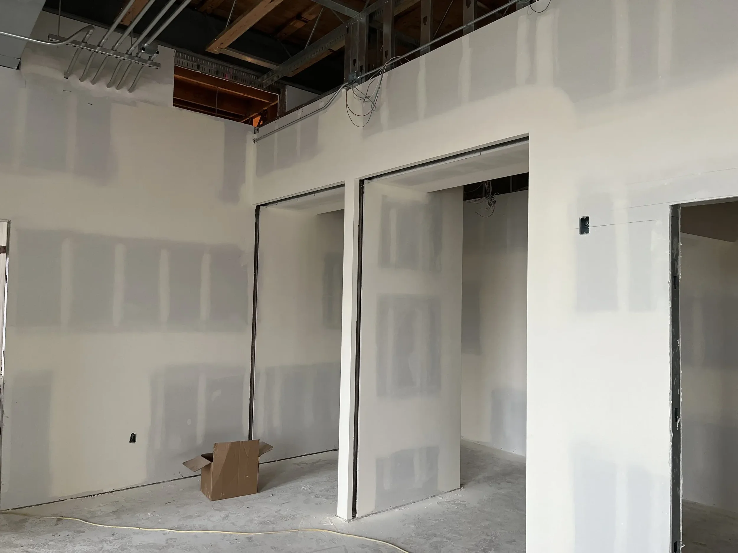 Interior of a building under construction with drywall installed on the walls, exposed ceiling with metal framing, electrical wiring hanging, and an open doorway.