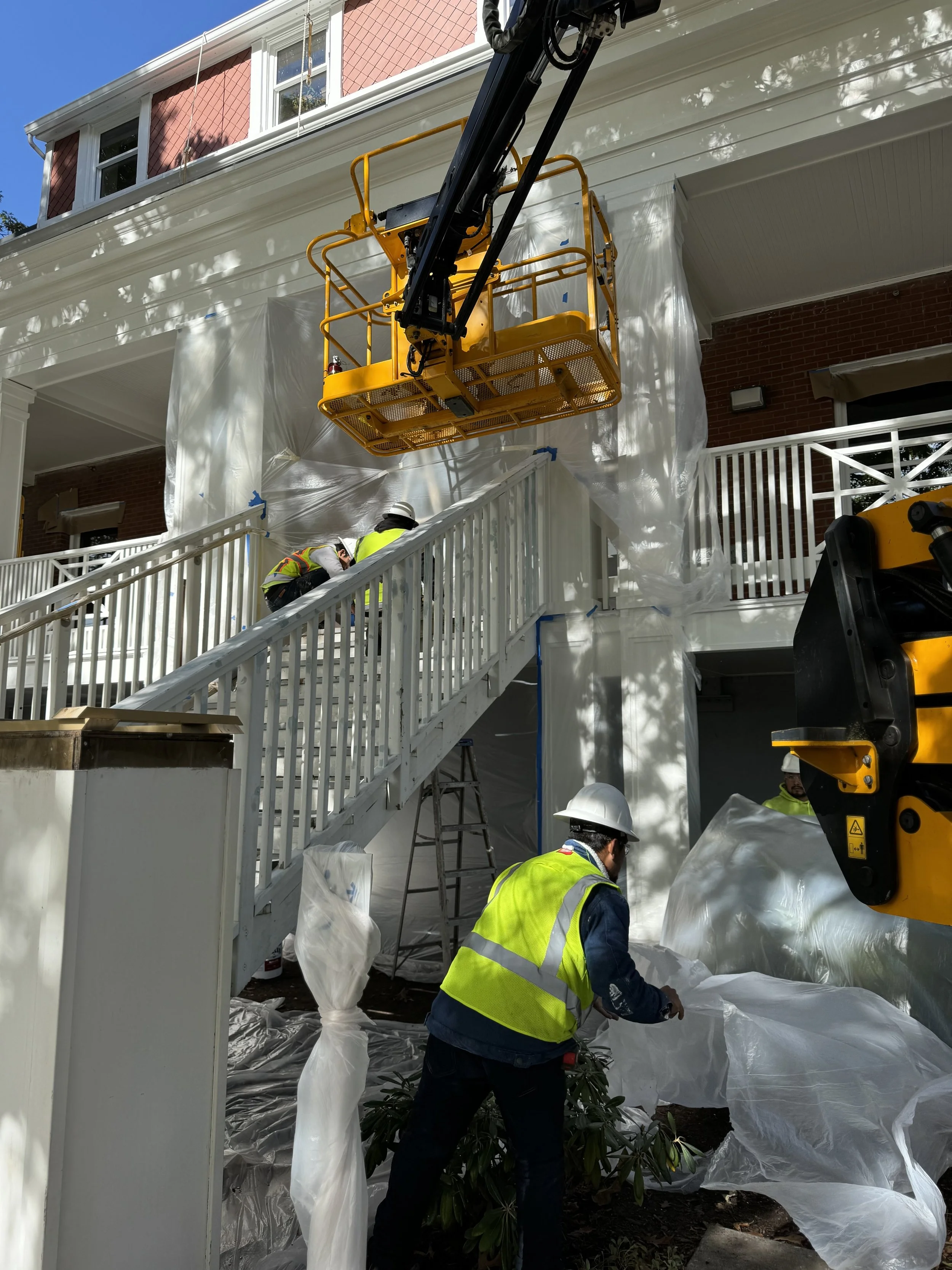 Construction workers in safety vests and helmets working outdoors on a building renovation, with a cherry picker lift elevated near the house, covered in white protective plastic sheeting.
