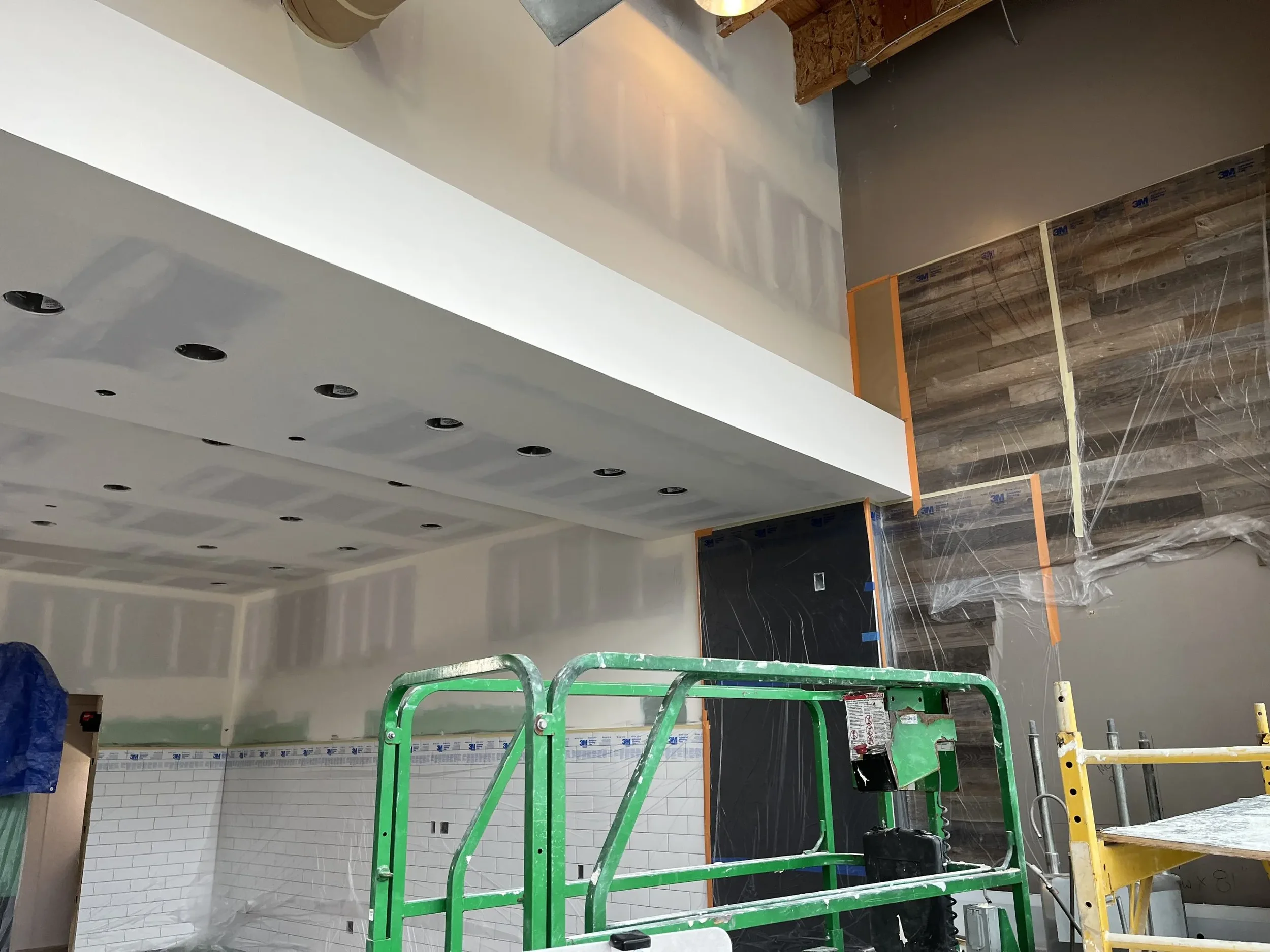 Interior of a construction site showing a white ceiling with recessed lighting holes, a partially finished wall with white brick tiles, and construction equipment including a green scaffold and supplies. The wall has a section with wood paneling and plastic sheeting.