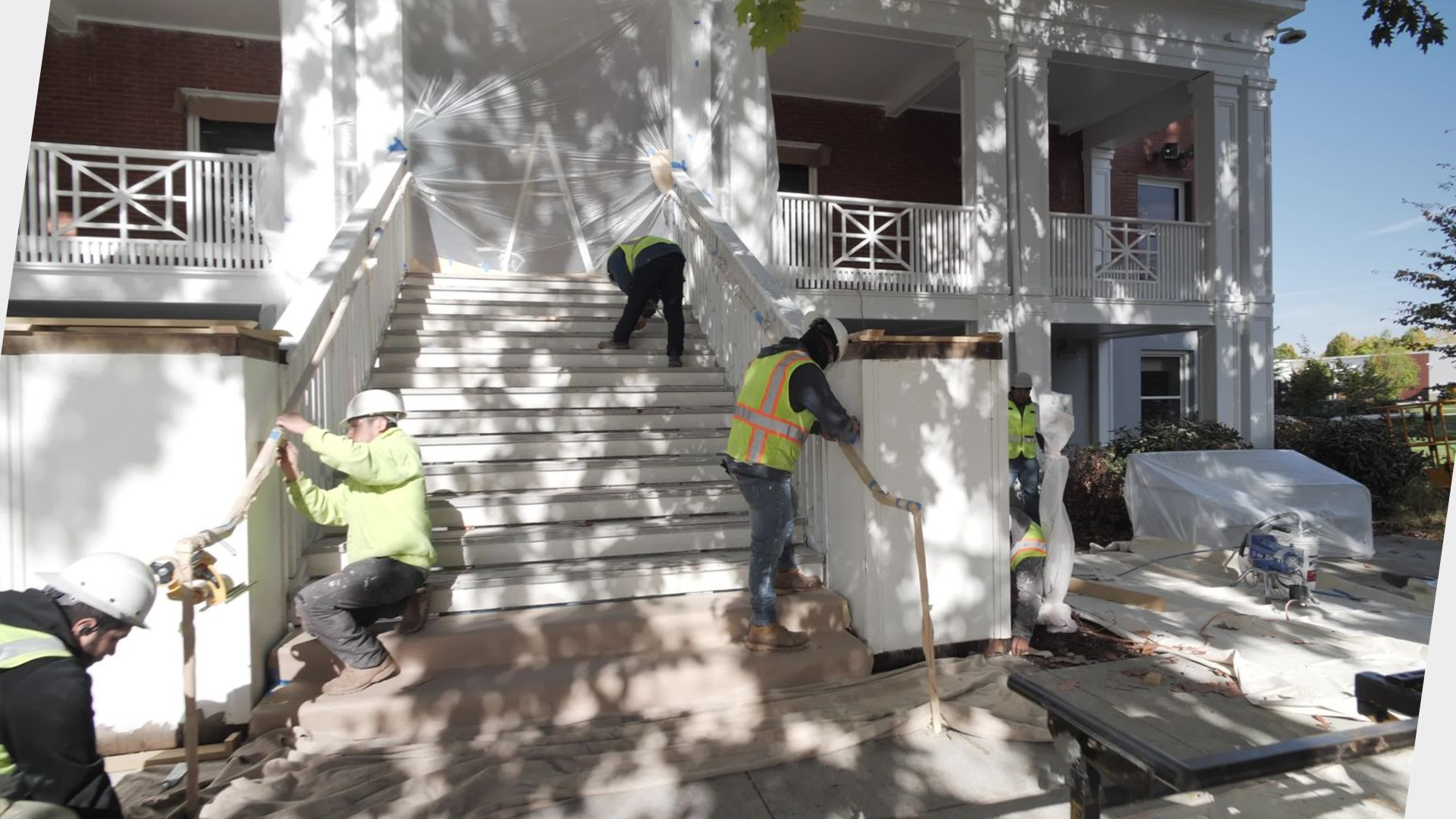 Construction workers installing or repairing white exterior stairs and railings of a house with a second-story porch, surrounded by shadows of trees on a sunny day.