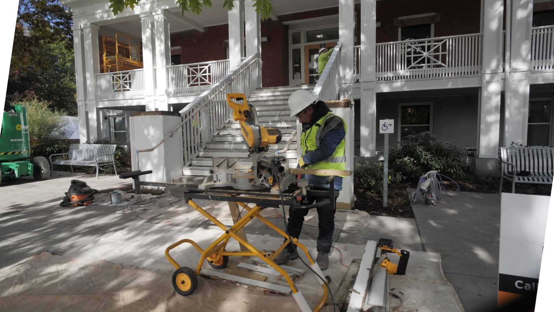 Construction workers installing or repairing stairs outside of a building. One worker in a yellow safety vest and white hard hat is operating a miter saw, while another worker in a yellow safety vest and white hard hat is working on the stairs. The setting includes white railings and columns, with trees and benches nearby.