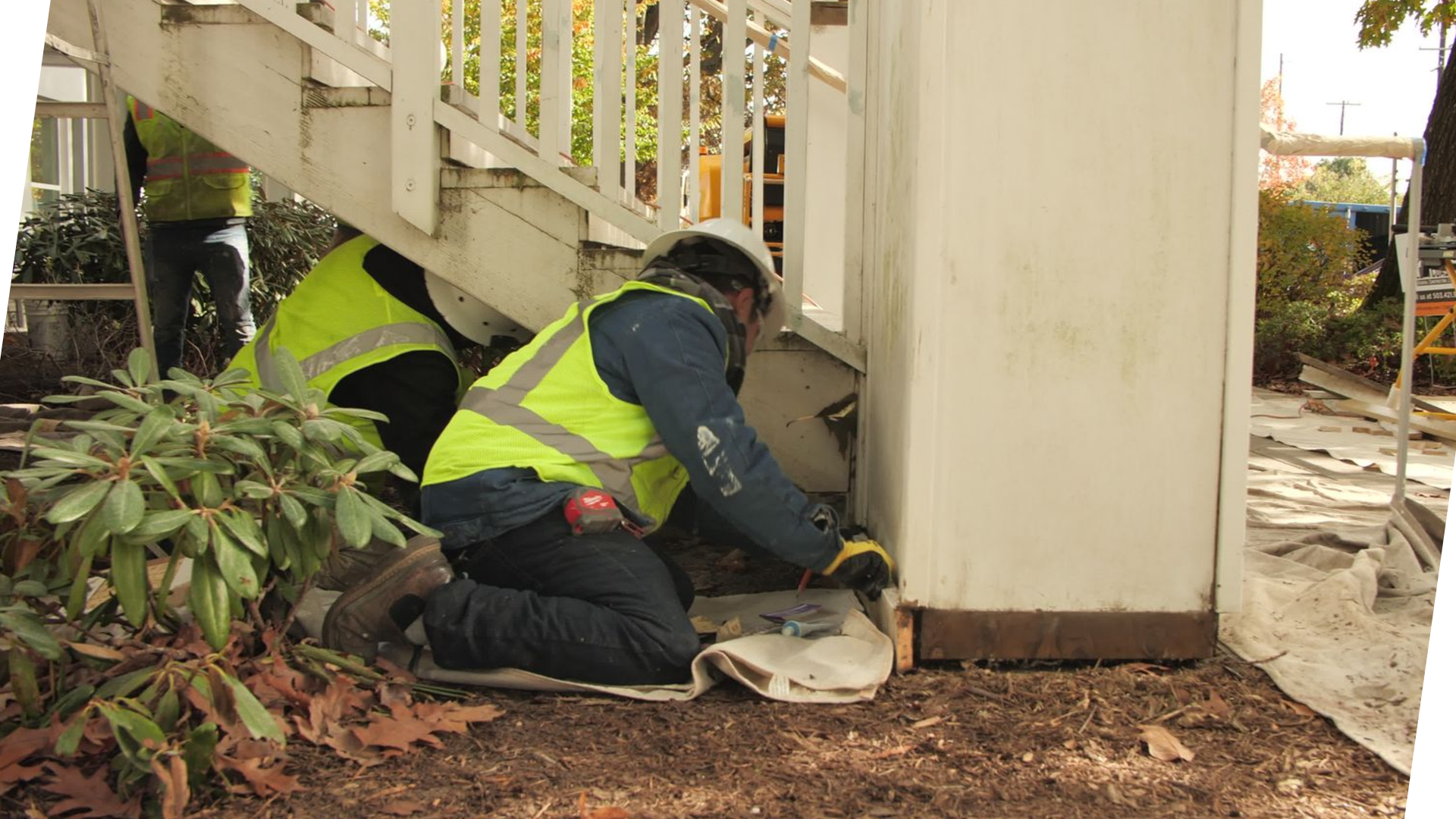 Two construction workers wearing safety vests and helmets kneeling under a wooden staircase, repairing or inspecting the foundation of a building outdoors.