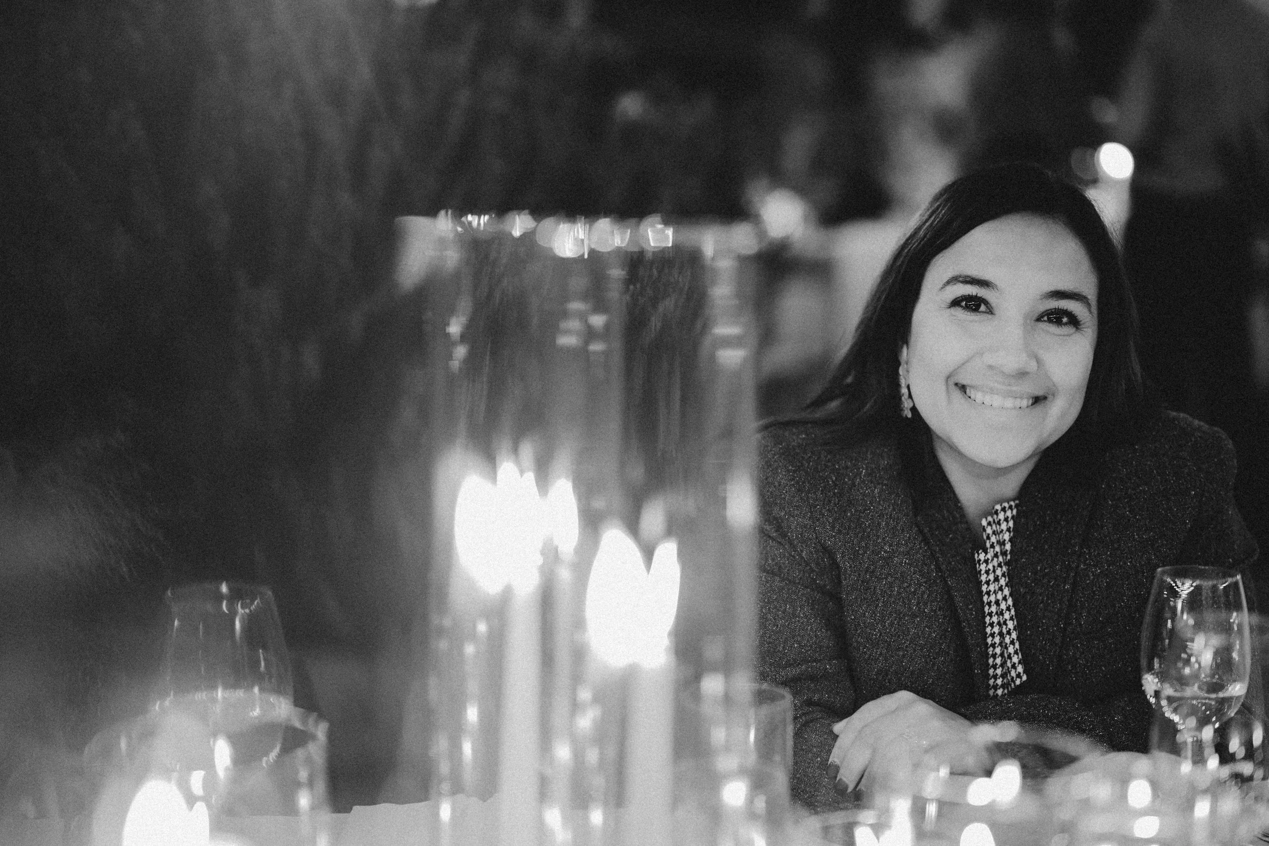 Black and white photo of a woman with dark hair smiling at a dining table at night. There are wine glasses and candles in the foreground.