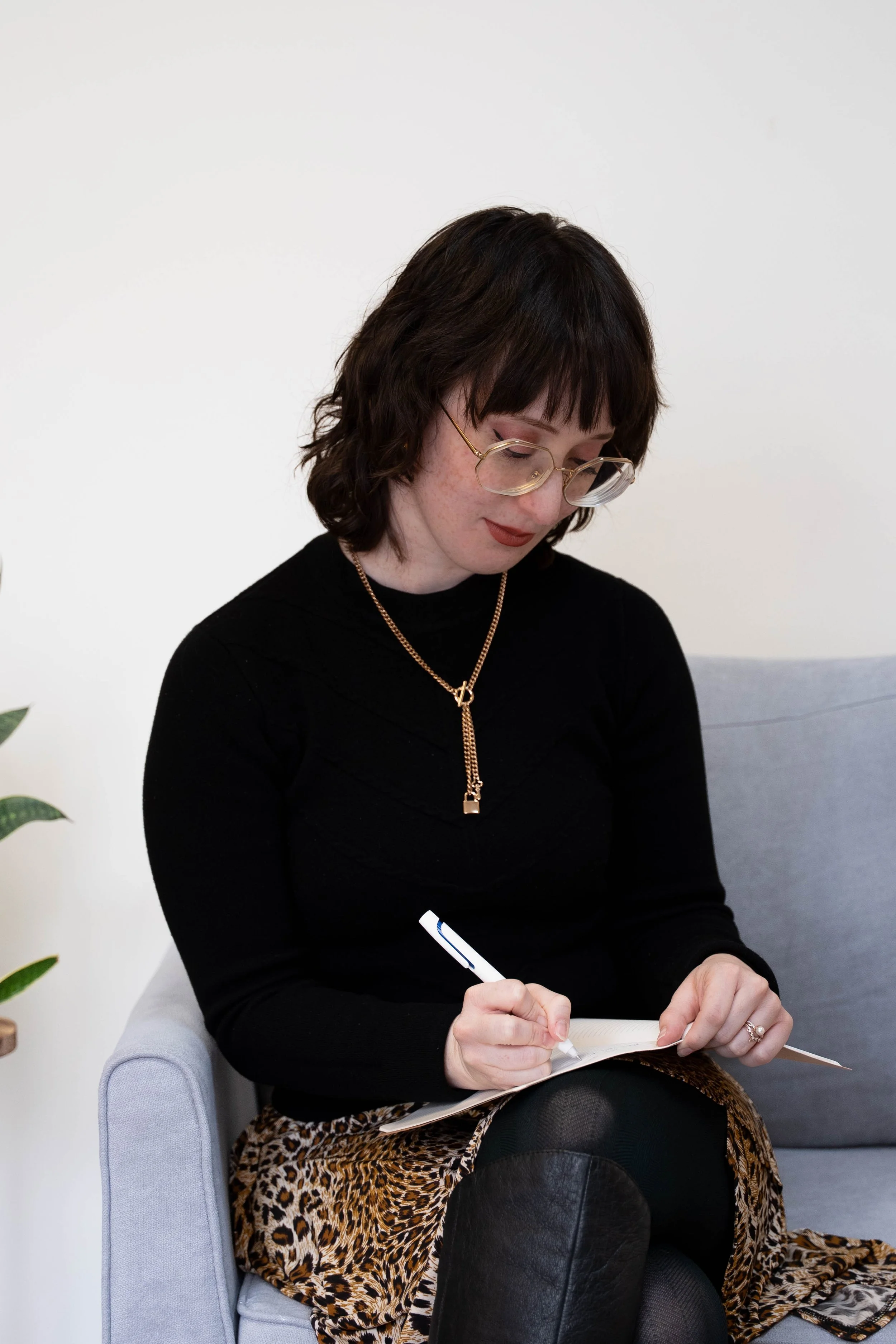 The therapist, a white woman with glasses in her late 20s, is centered in the photograph seated on the right side of the couch. Her head is tilted down slightly and she is writing something in a notebook, looking focused.
