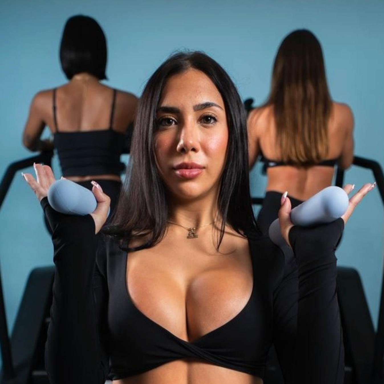 Woman with dark hair and a black workout top holding gray dumbbells, with two women in black workout clothing in the background at a fitness gym.