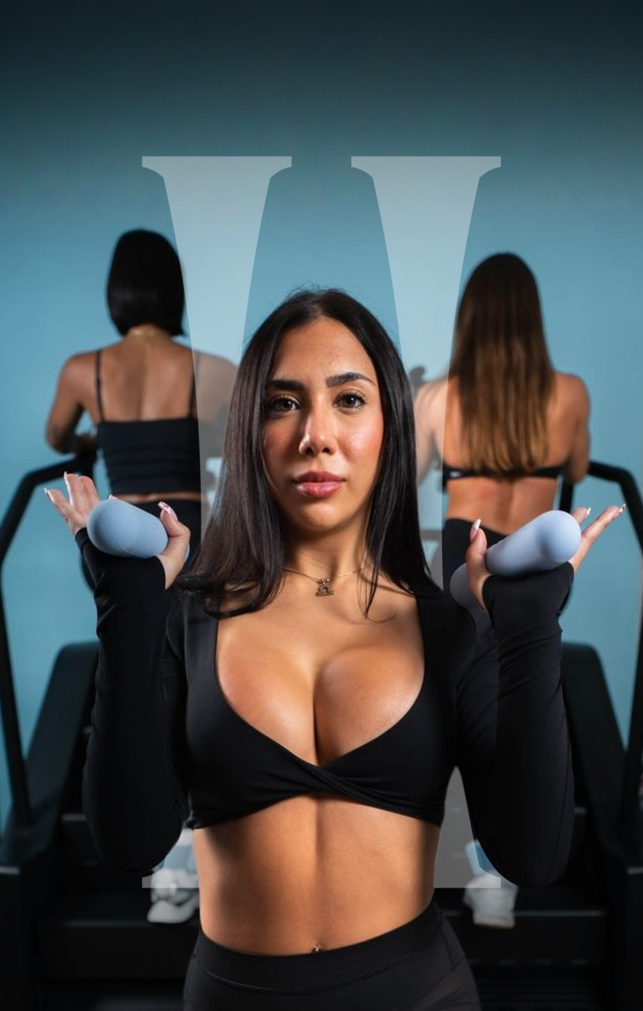 A woman with long dark hair, wearing a black crop top and black leggings, sits with her arms outstretched holding yoga stones, in front of two women doing yoga in a gym or studio. The studio wall features a large, blurred Hourglass in the background.