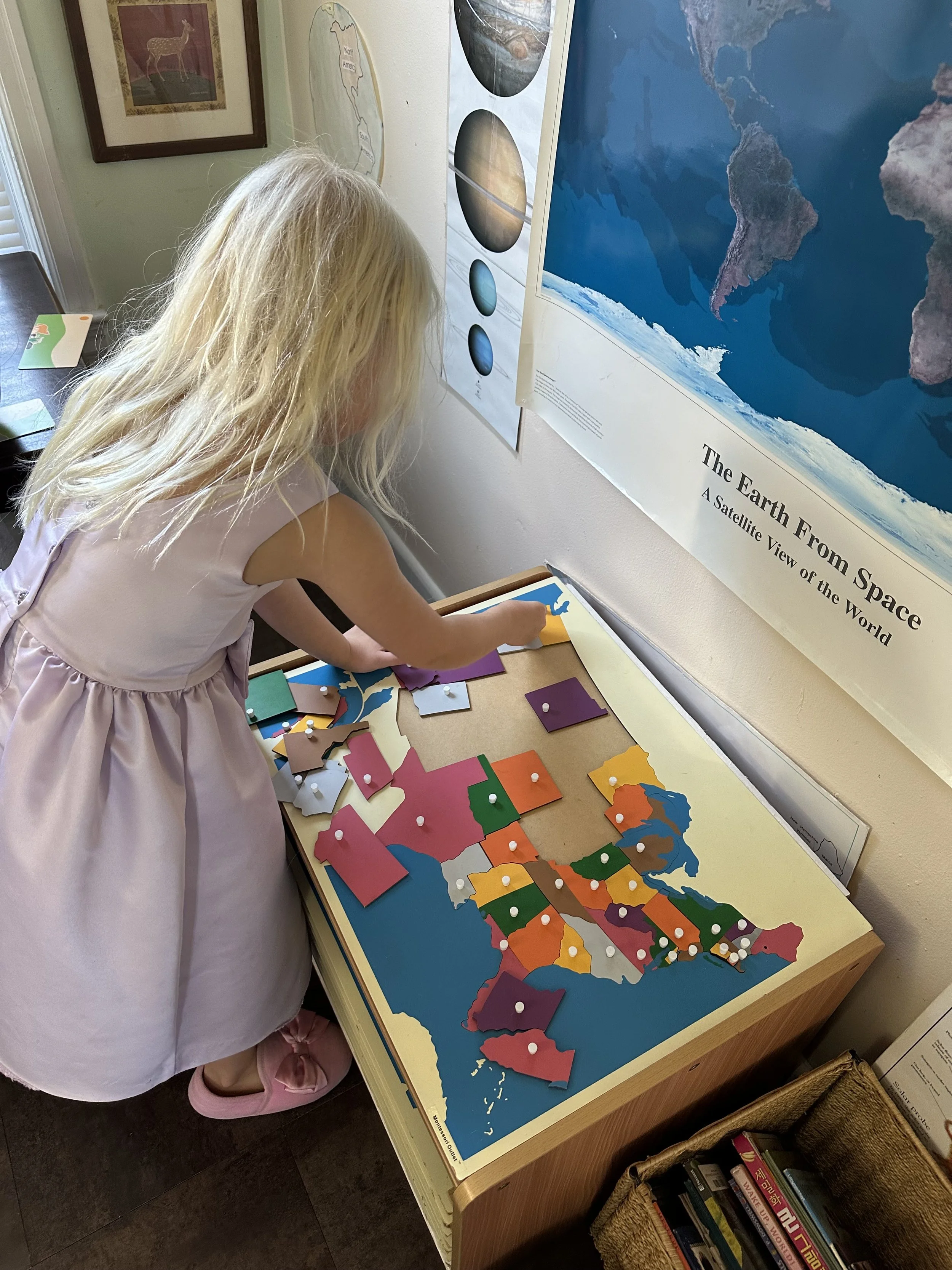 A young girl in a pink dress and pink shoes is standing at a table, placing a puzzle piece onto a map of the United States, which is colored by state. There are educational posters on the wall, including one about space and Earth.