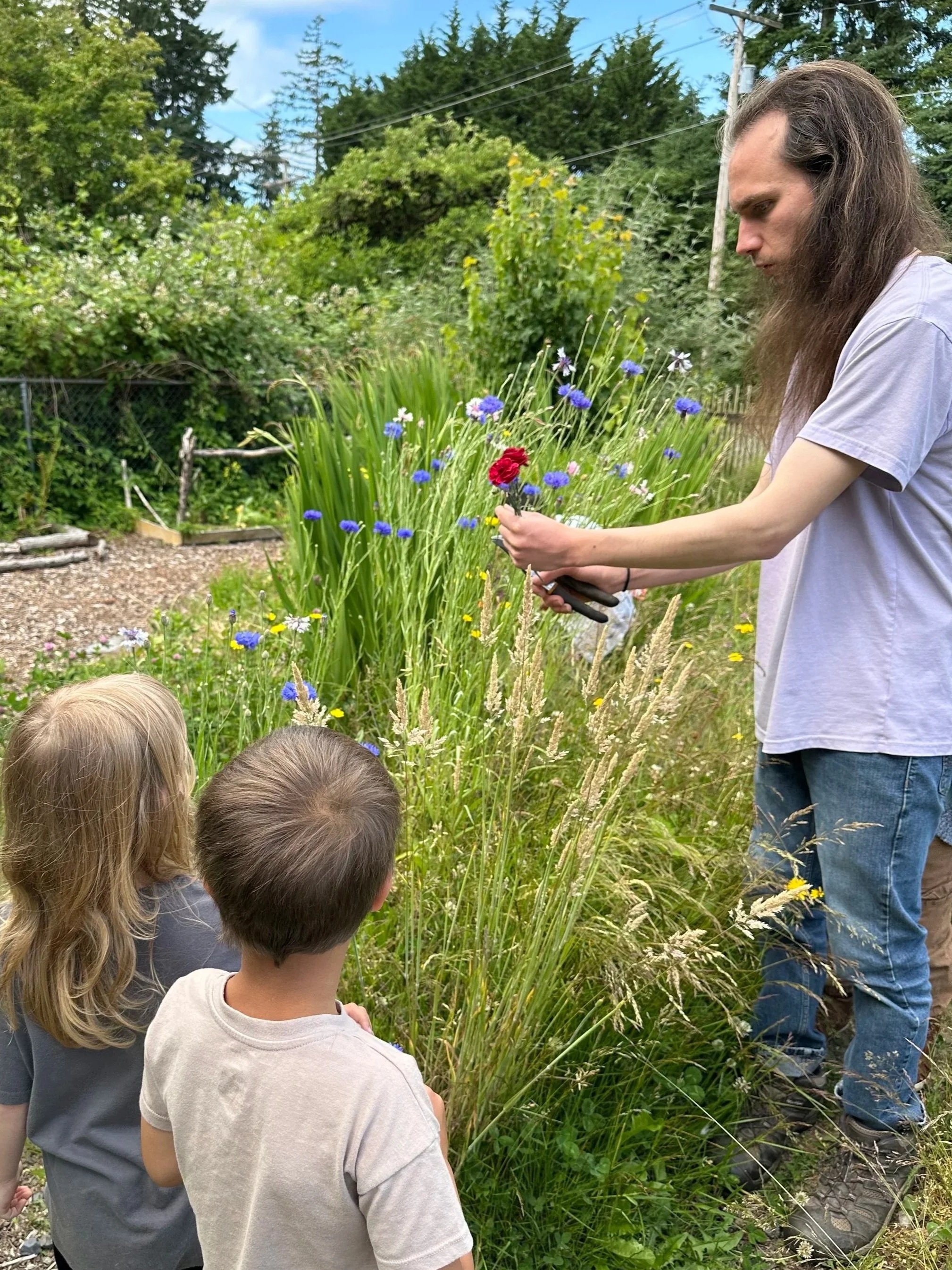 A man shows a bunch of colorful flowers to two children in a garden with various plants and trees.