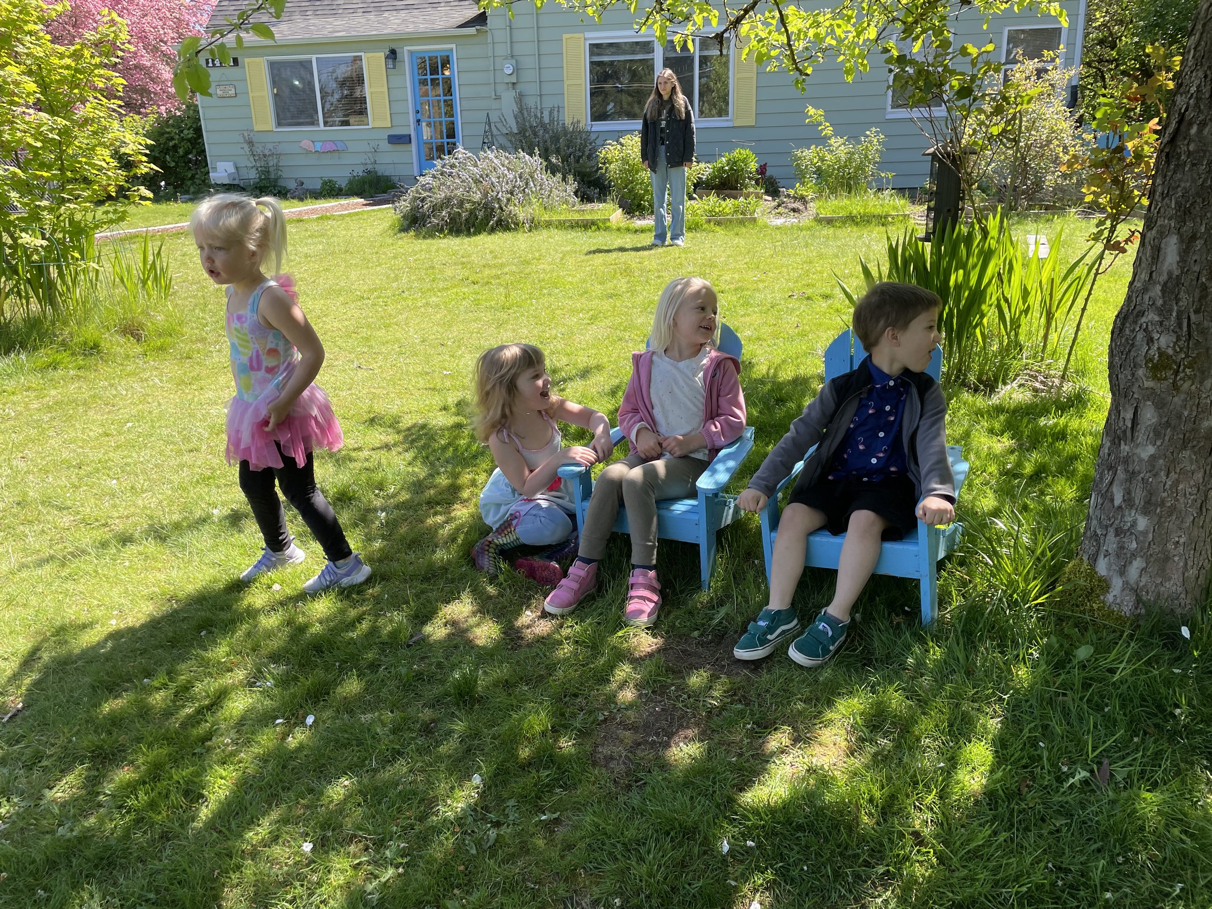 Four children playing outdoors on a sunny day with a house in the background. Three children are sitting on blue chairs under a tree, and one girl is standing nearby, wearing a colorful top and black pants.