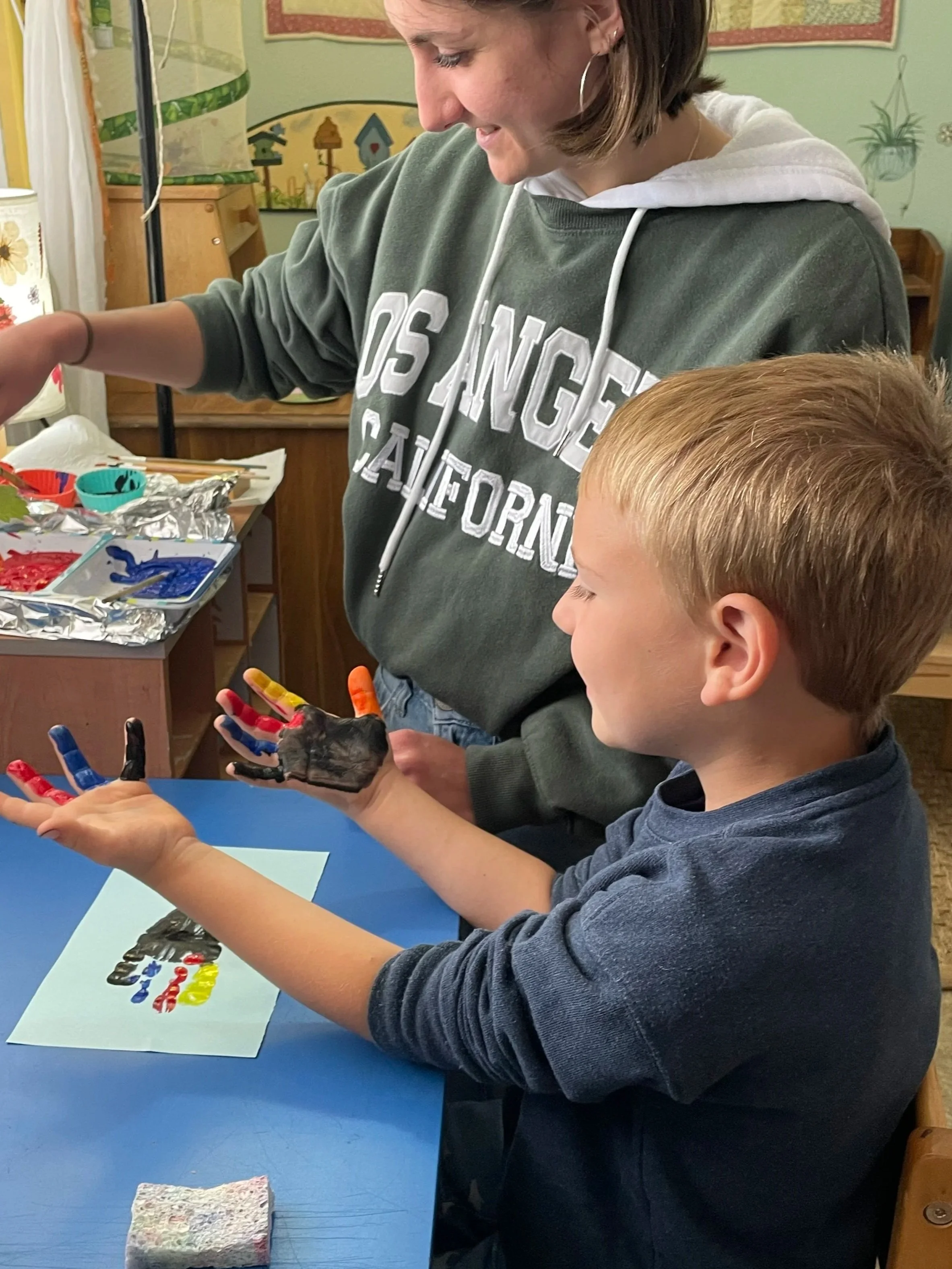 A young boy with painted hands sitting at a blue table, showing a colorful paint project, with a woman standing beside him smiling.