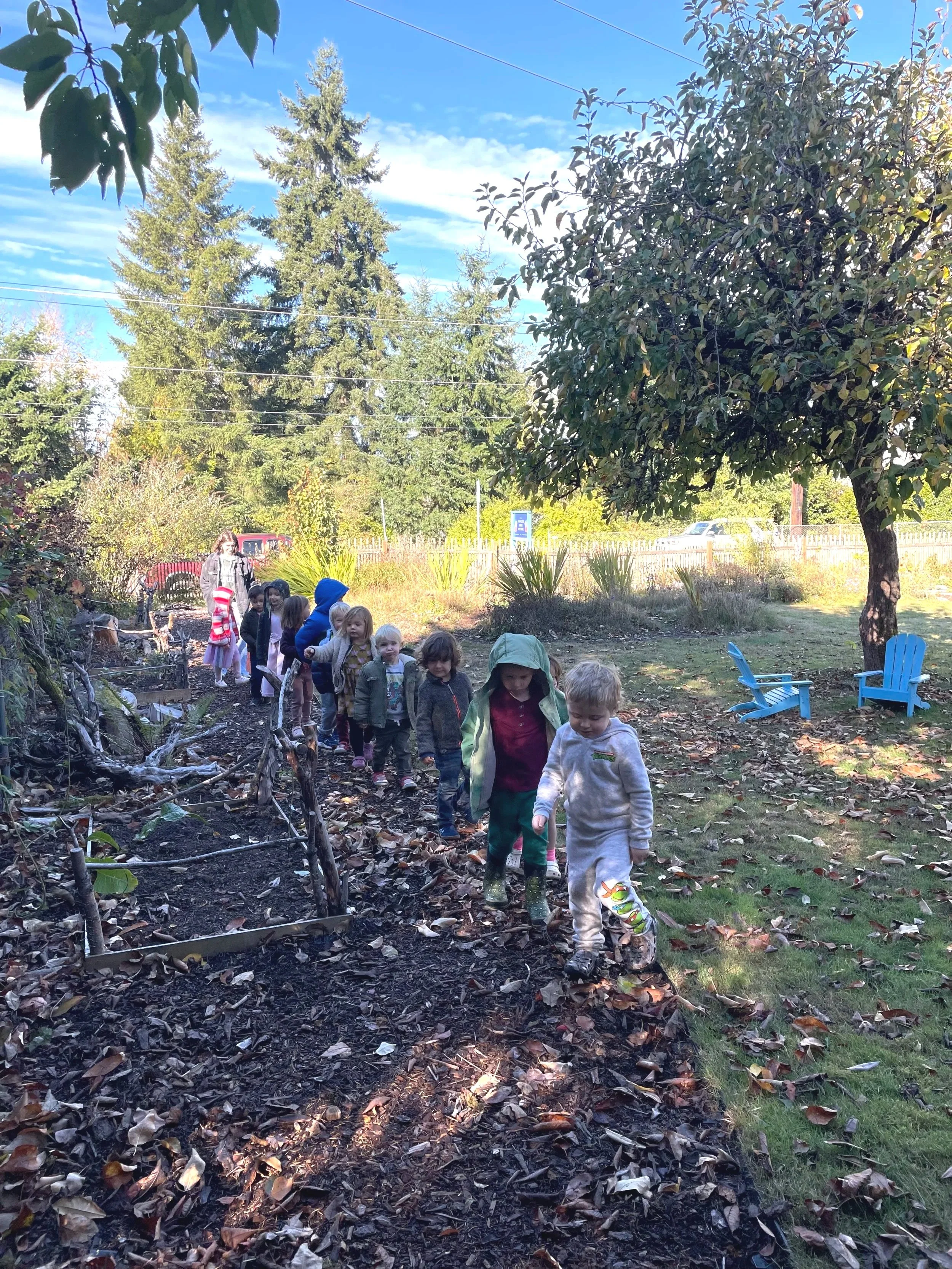 Children walking in a line on a dirt path in a garden with trees and bushes, some adult supervision, blue sky with some clouds visible.