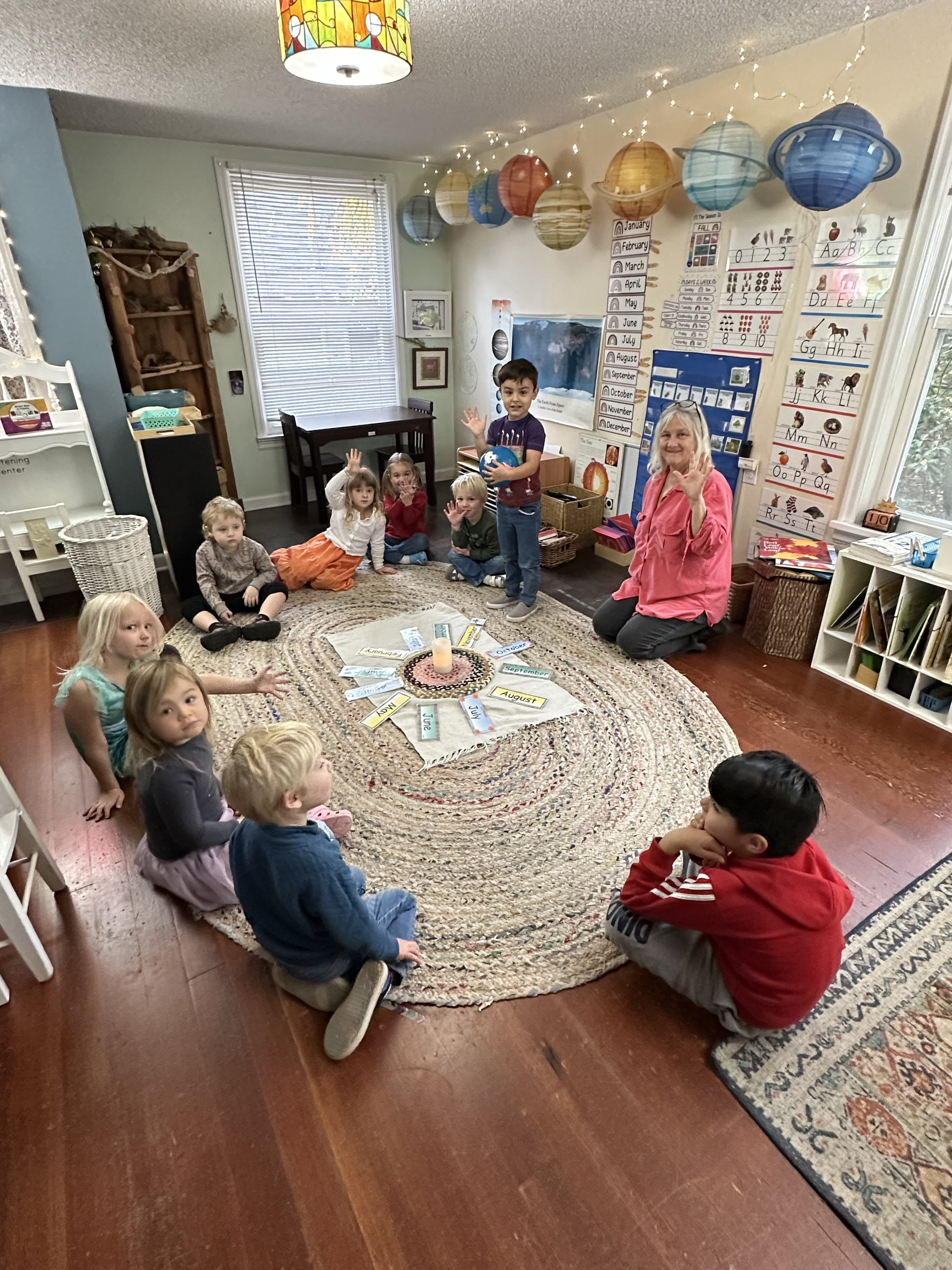 Children and a teacher sitting in a circle in a classroom, engaging in an activity around a center decorated with seasonal and month labels, with educational posters and decorations on the walls and a window in the background.