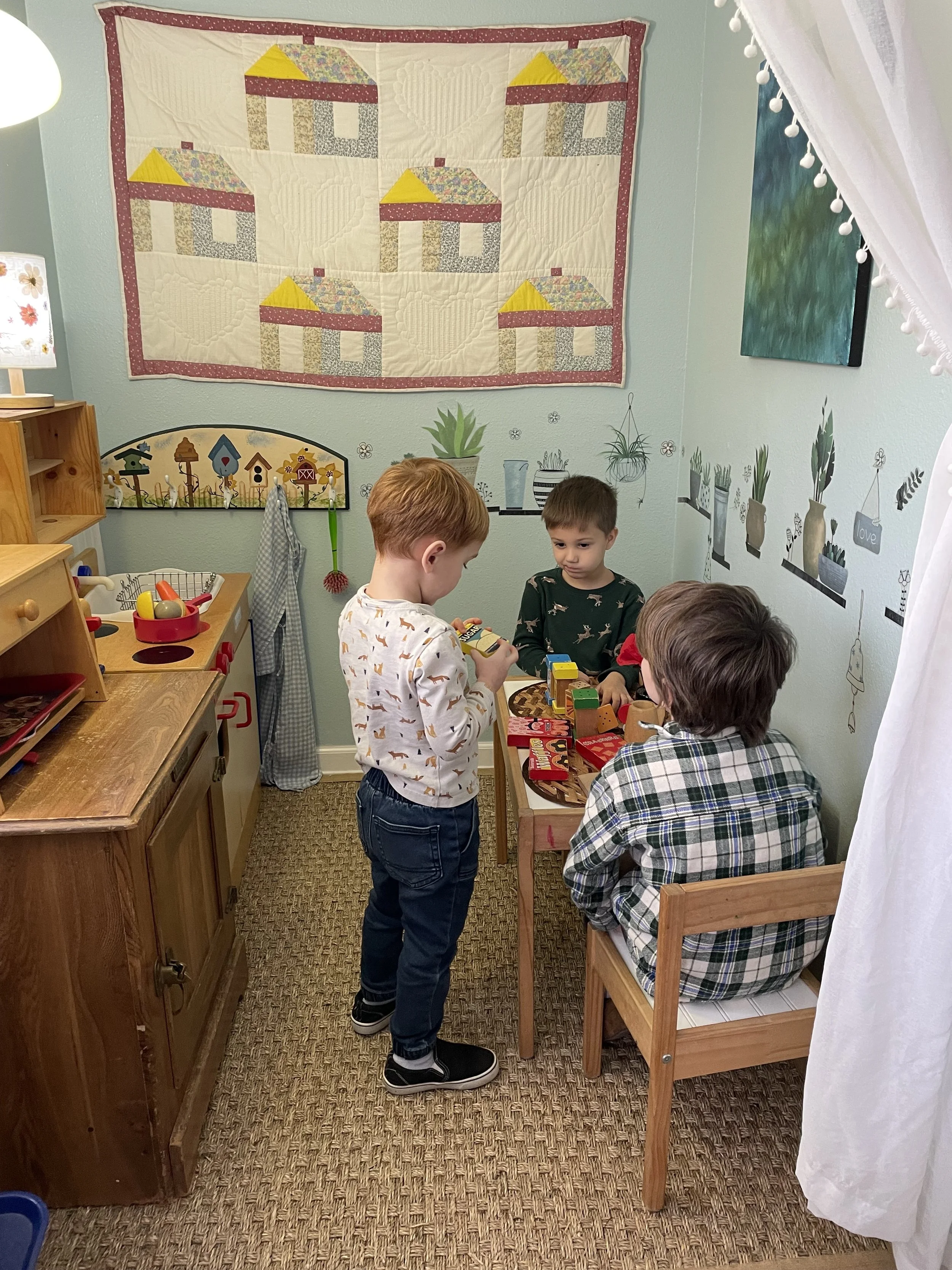 Three children playing with wooden toy blocks in a kitchen-themed playroom.