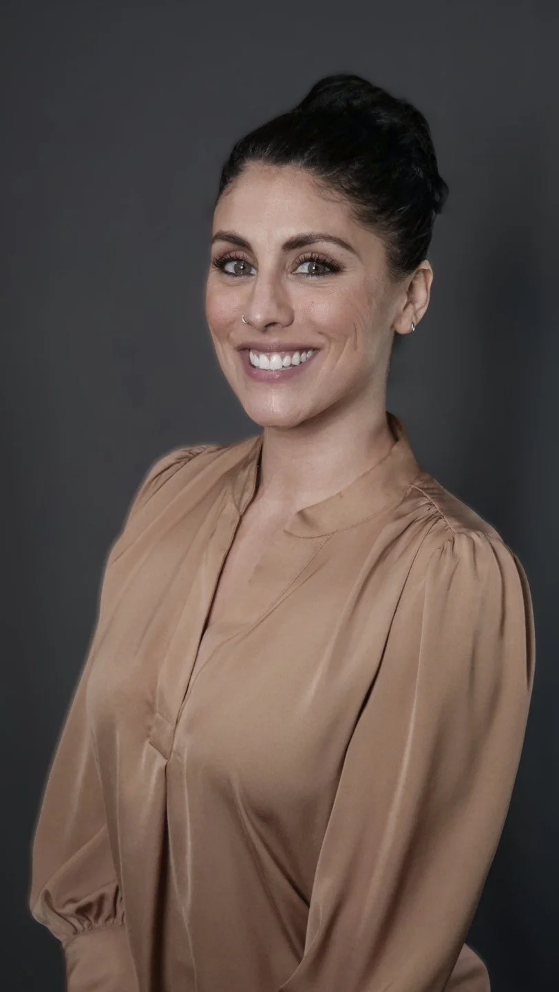 A woman with dark hair styled in an updo, smiling, wearing a tan blouse, against a dark gray background.