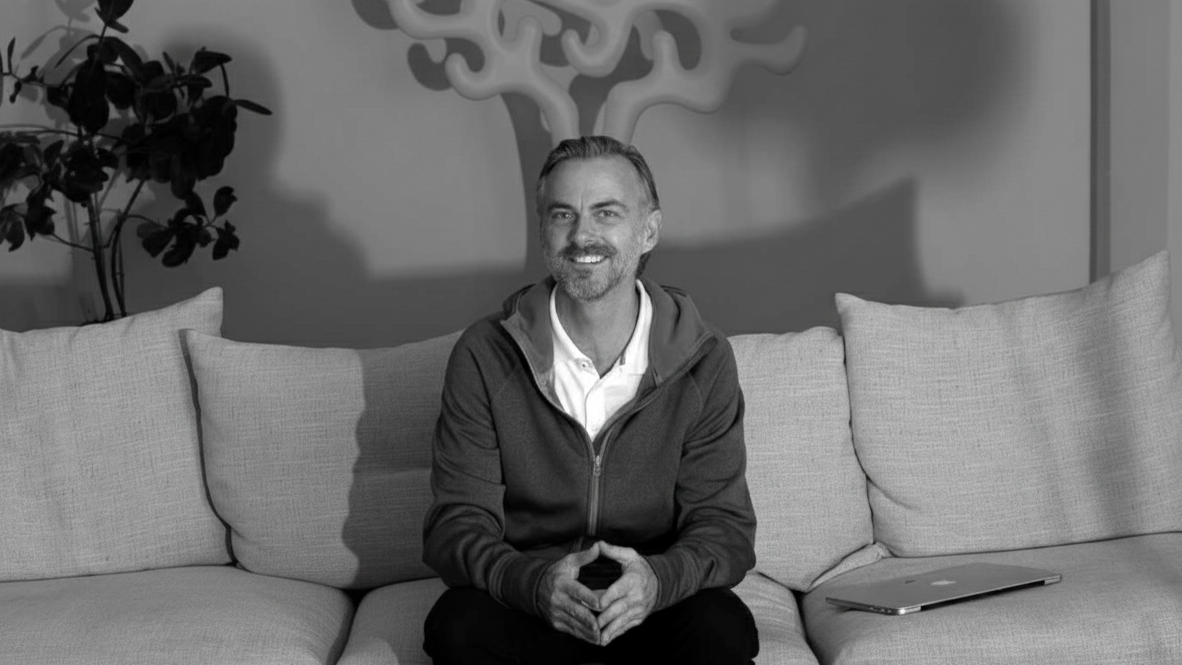 Gareth Stapleton | A man sitting on a light-colored sofa with cushions, smiling, in a room with a decorative wall piece and a plant in the background, in black and white
