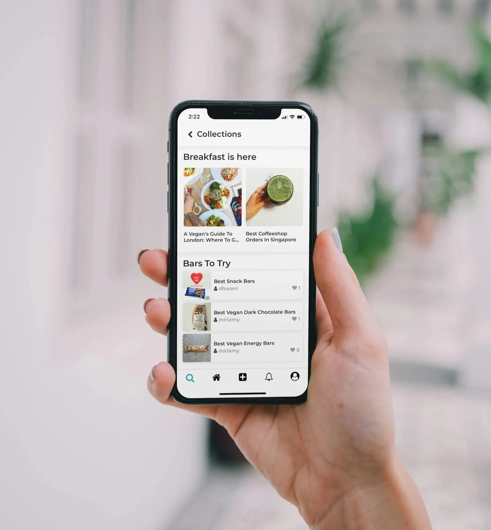 A person holding a smartphone displaying a food app with sections for breakfast collections and vegan snack bars, with plants and a window in the background.