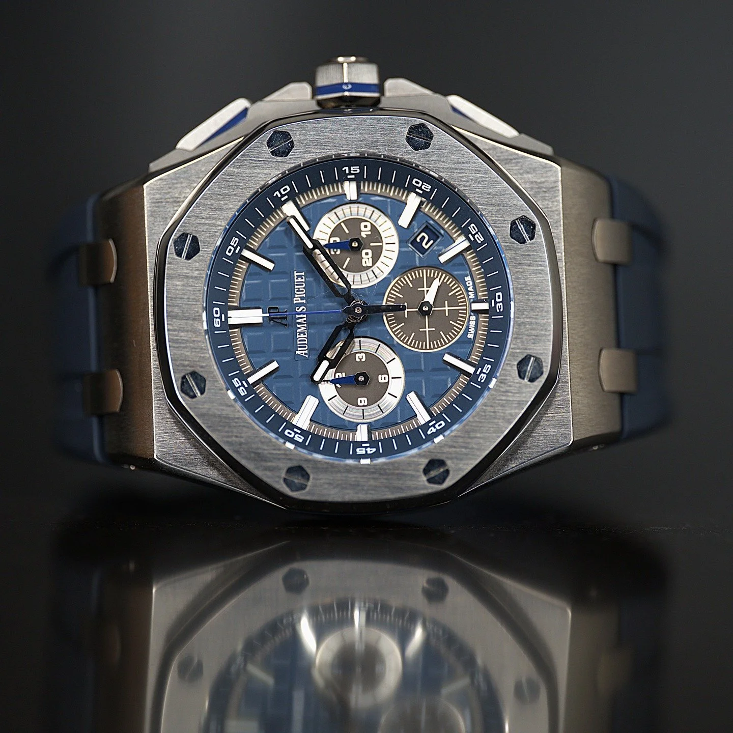 A luxury wristwatch with a blue dial, chronograph subdials, and a brushed stainless steel bezel, resting on a reflective surface.