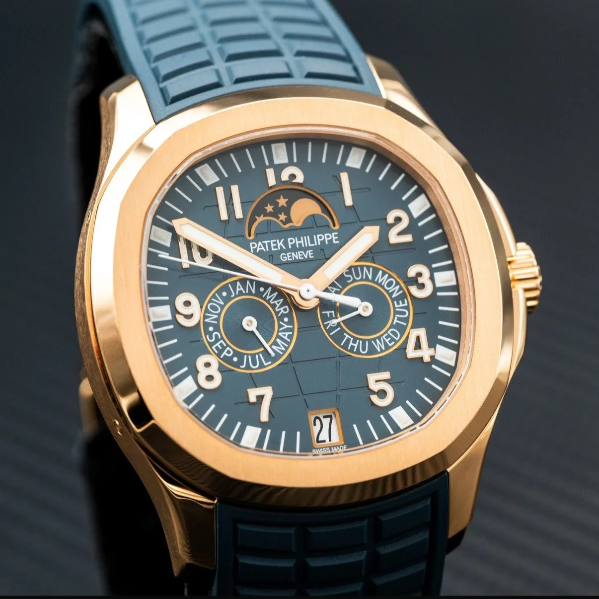 A luxury Patek Philippe wristwatch with a blue dial, gold case, and a blue strap. The watch displays multiple subdials for days, months, and a moon phase indicator, along with a date window at the 6 o'clock position.