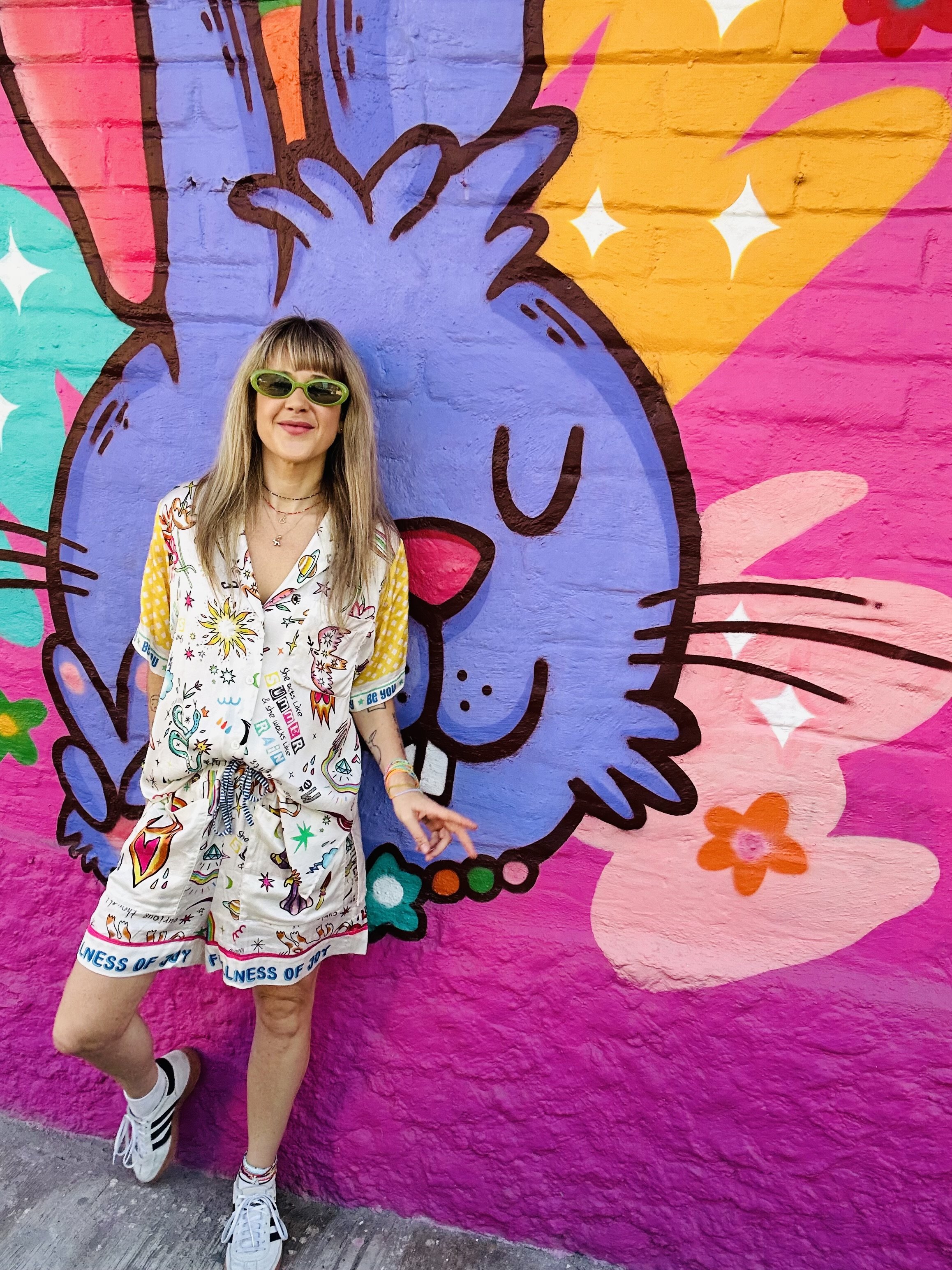 A woman with blonde hair, wearing green sunglasses, colorful printed matching shirt and shorts, standing in front of vibrant street art of a blue cartoon-like animal face with pink, orange, and yellow background.