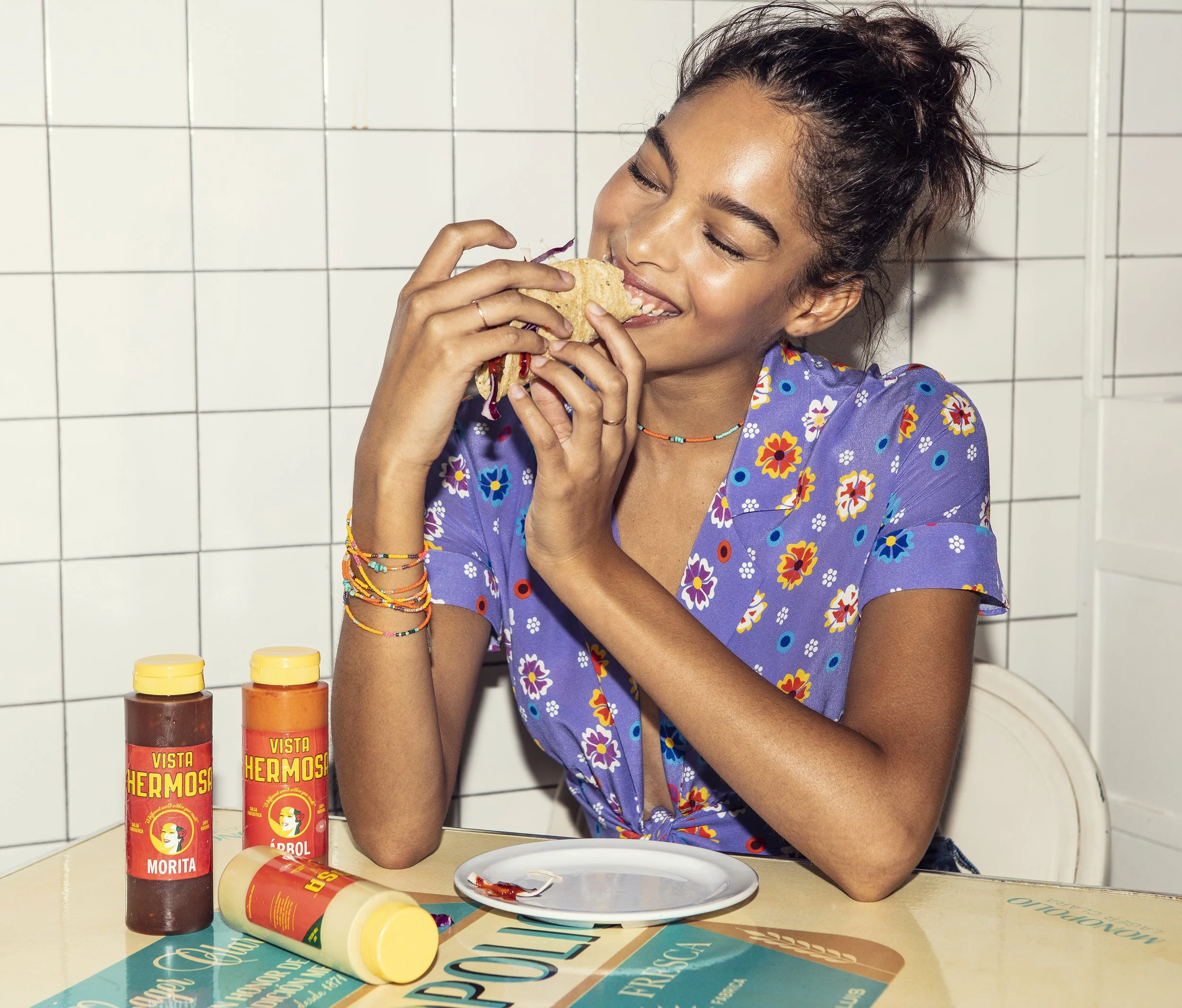 A woman in a floral purple LHD shirt enjoying a taco with bottles of hot sauce on the table in a kitchen with white tiled wall wearing colorful Bunny Shapiro bracelets.