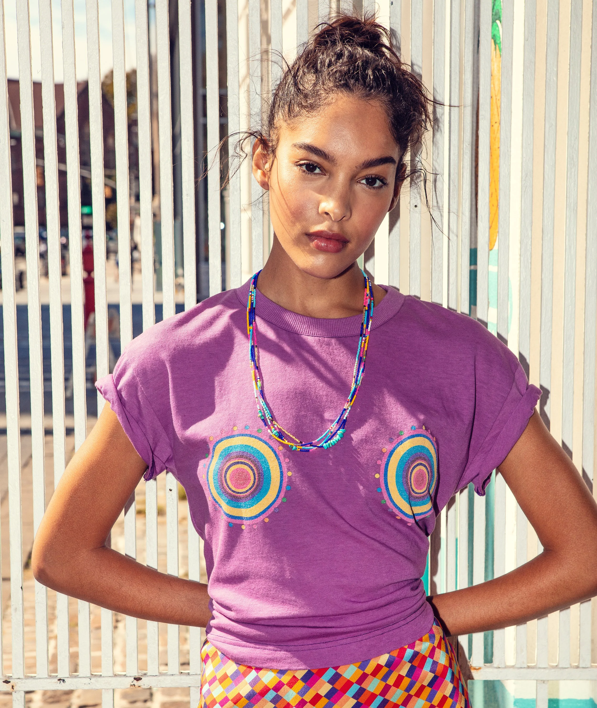Young woman with curly hair in a ponytail, wearing a purple graphic LHD T-shirt with colorful concentric circle designs and multicolored checkered pants, standing outdoors against a metal fence with vertical bars wearing Bunny Shapiro necklaces. 