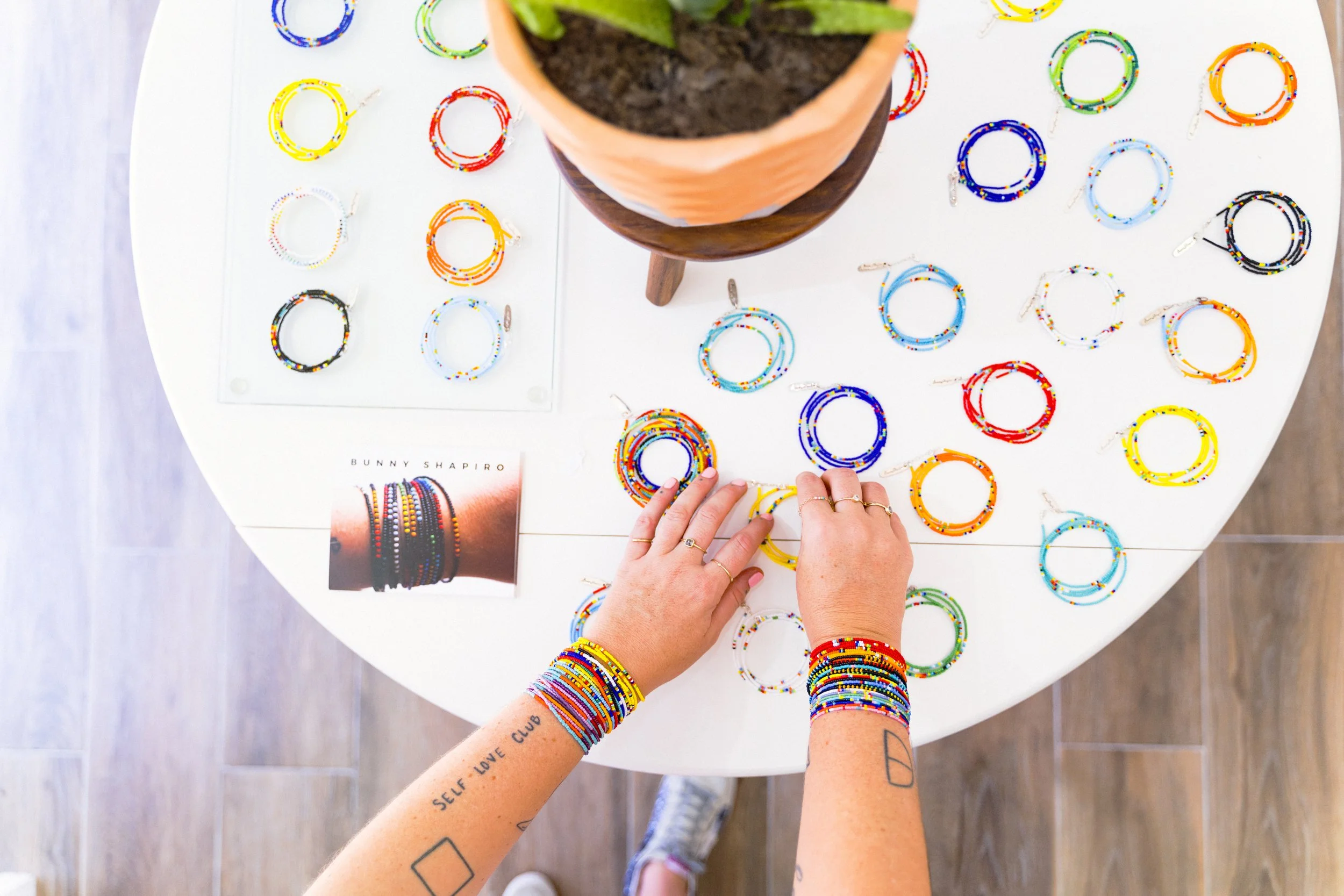 Person arranging colorful beaded bracelets on a white table, with a potted plant in the center and a display of bracelets in the background.