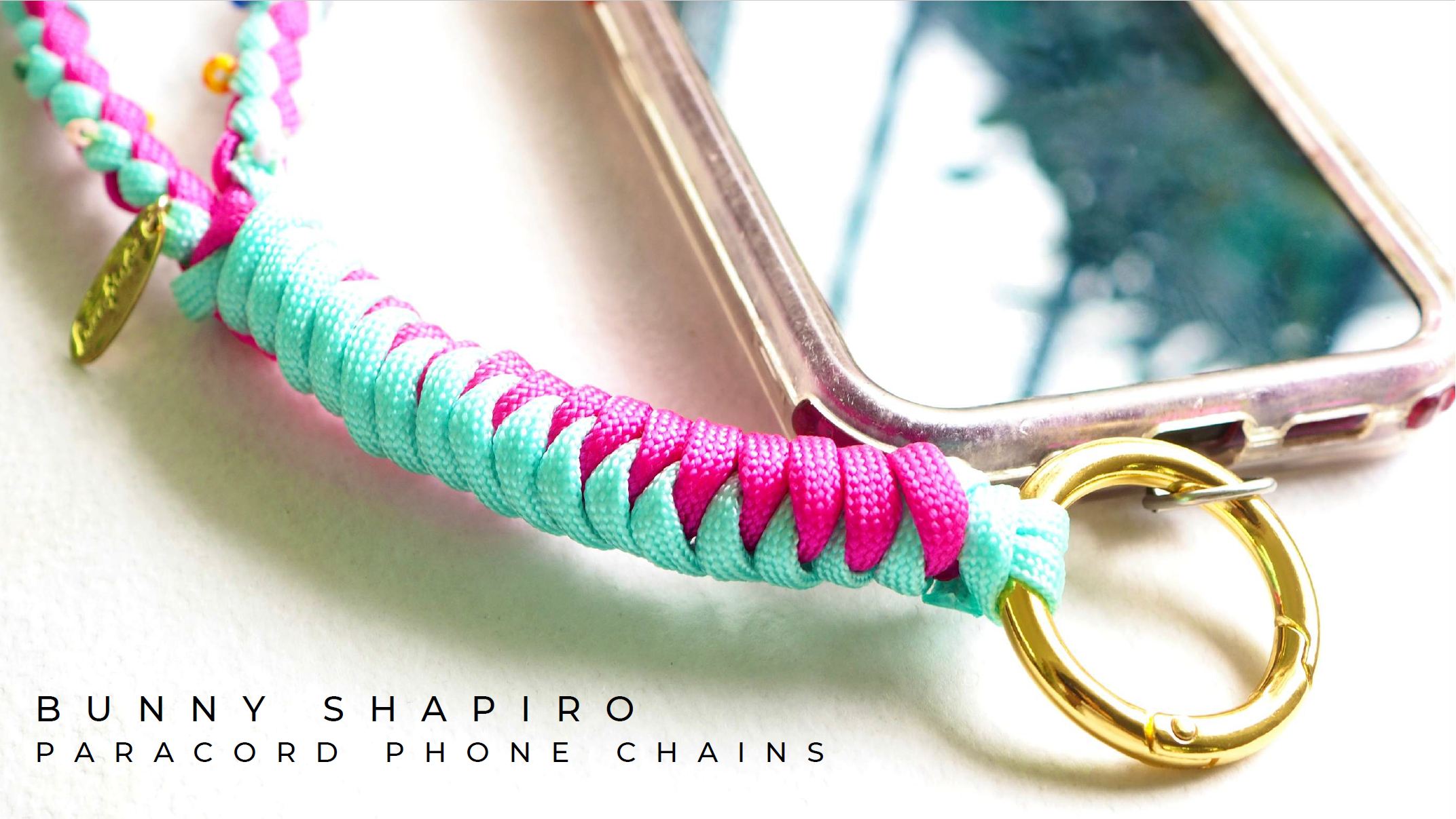 Colorful paracord phone chain in pink and turquoise attached to a pink phone case and a gold keyring, with text reading "Bunny Shapiro Paracord Phone Chains."