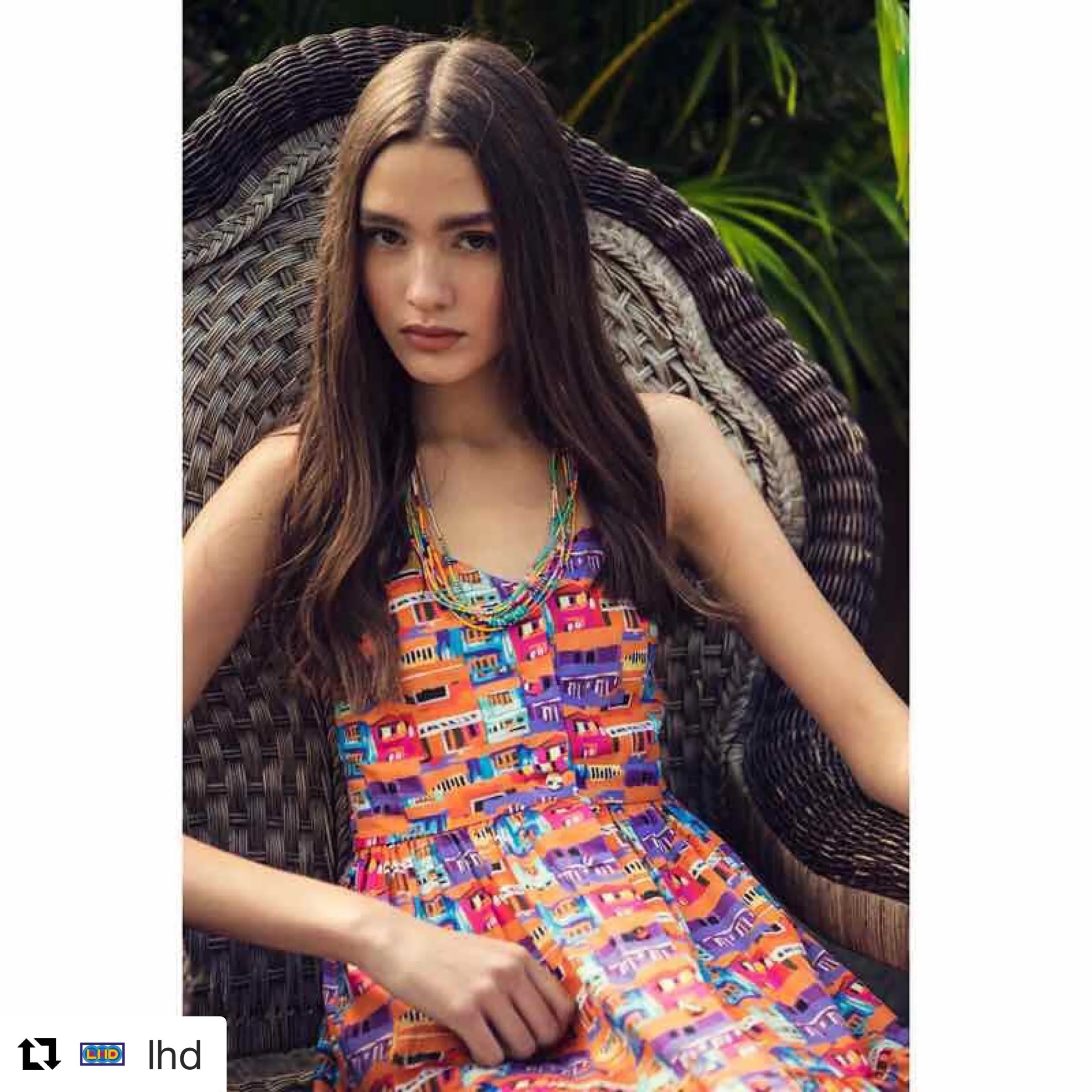 A woman with long brown hair sitting on a wicker chair surrounded by green foliage, wearing a colorful LHD sleeveless dress with a geometric pattern and layered Bunny Shapiro necklaces.