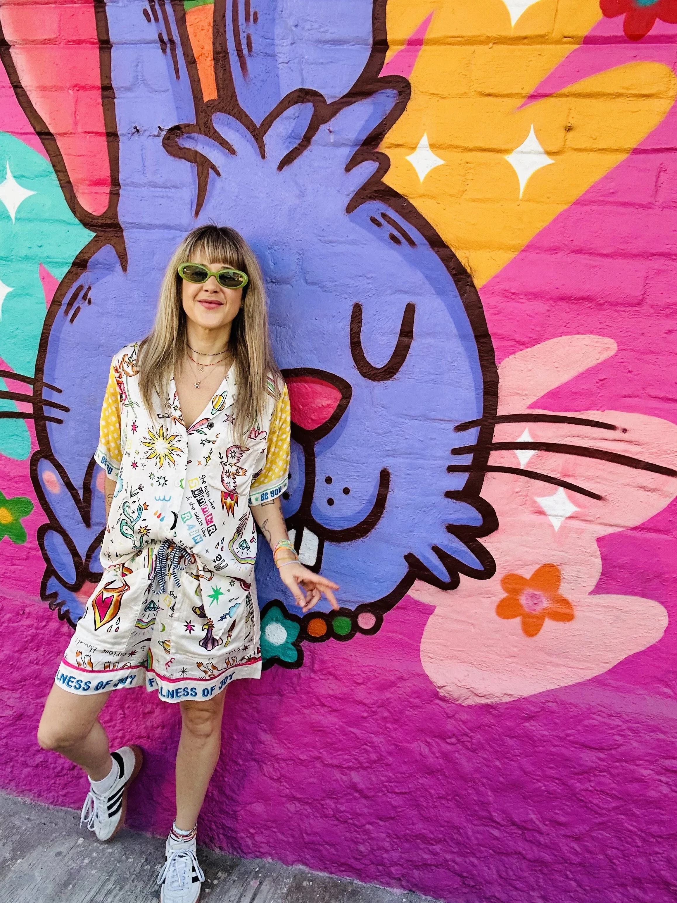 A woman with blonde hair wearing yellow sunglasses, a colorful printed shirt, and matching shorts stands in front of a vibrant mural of a blue cartoon cat with pink, yellow, and orange background elements.