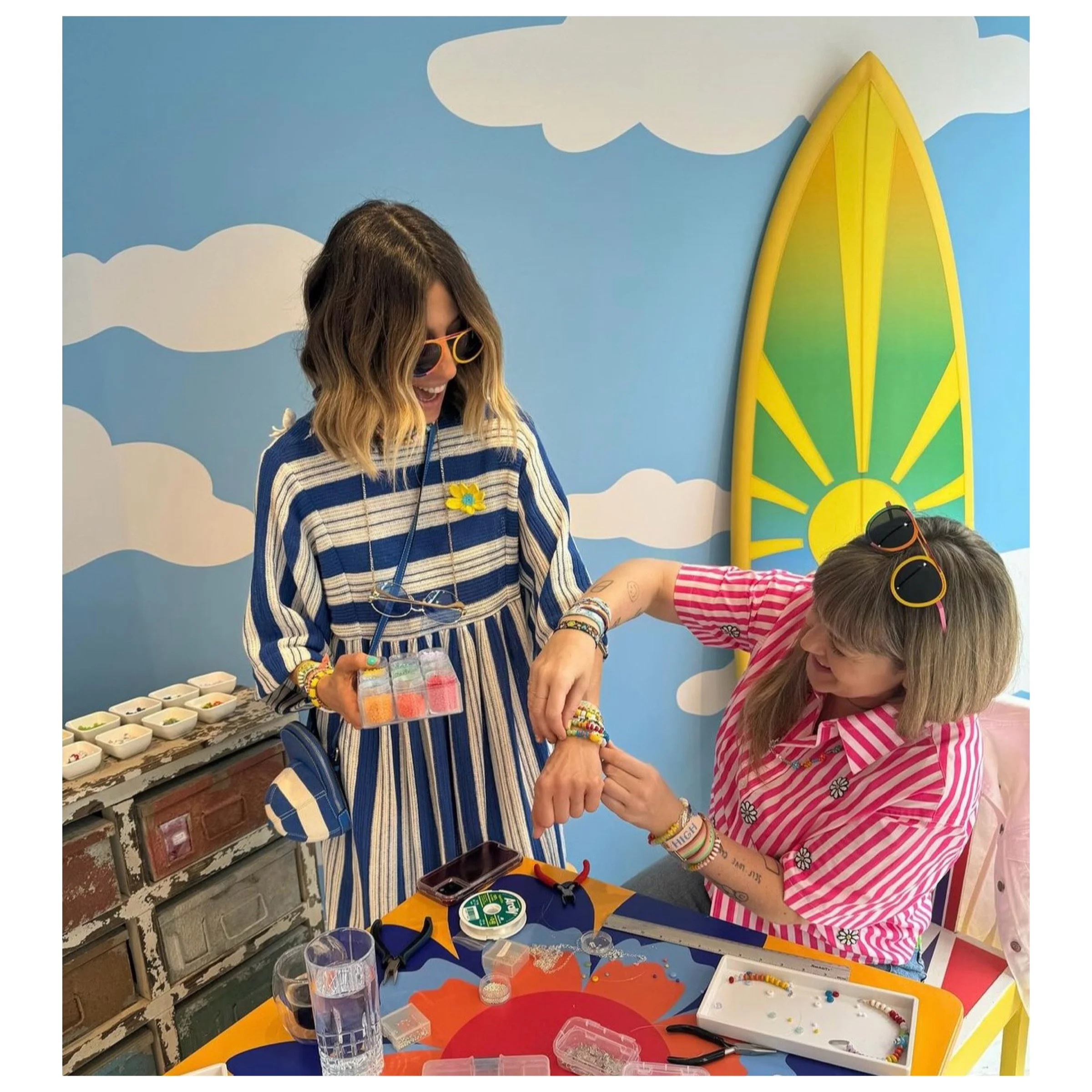 Bunny Shapiro and Mira Mikati with colorful bracelets and sunglasses are smiling while creating jewelry at the Happy House in London UK. The background features a painted sky with clouds and a large yellow and green surfboard.