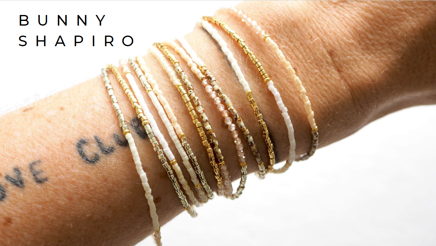 Various delicate beaded bracelets in white, gold, and pink tones on a person's wrist, with a tattoo partially visible, against a white background.