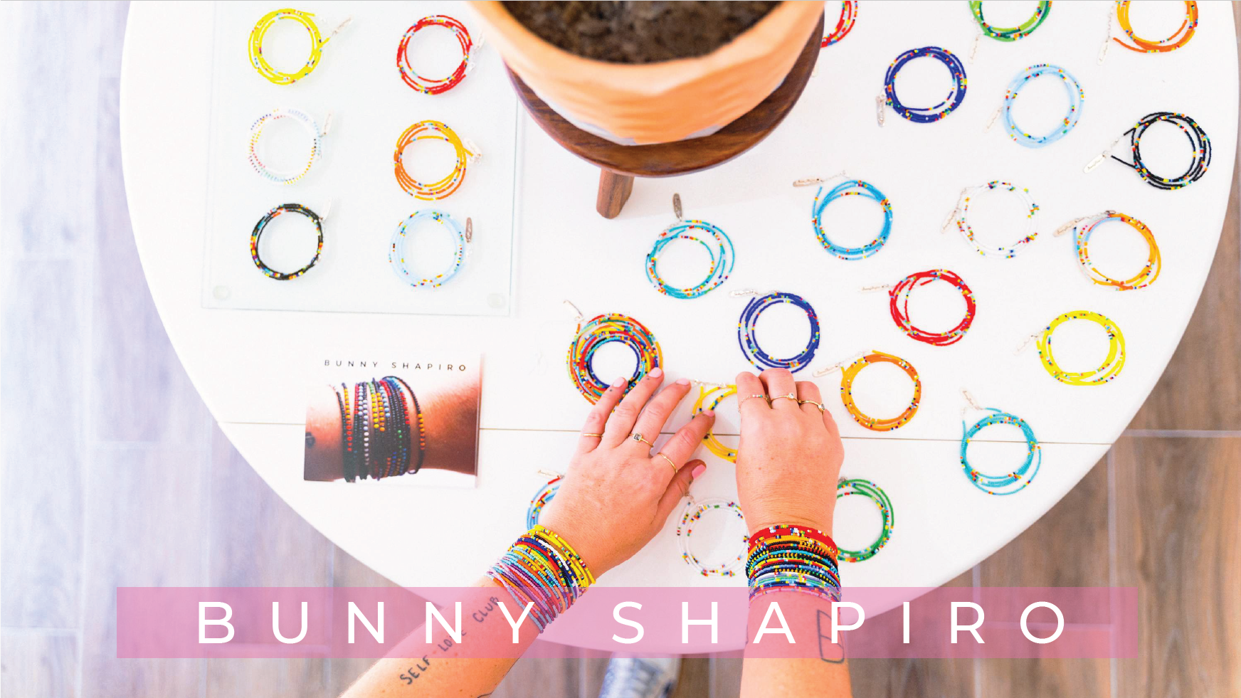 Person arranging colorful beaded jewelry bracelets on a white table with a display card and a plant on a wooden stand. The text 'BUNNY SHAPIRO' is overlaid at the bottom of the image.