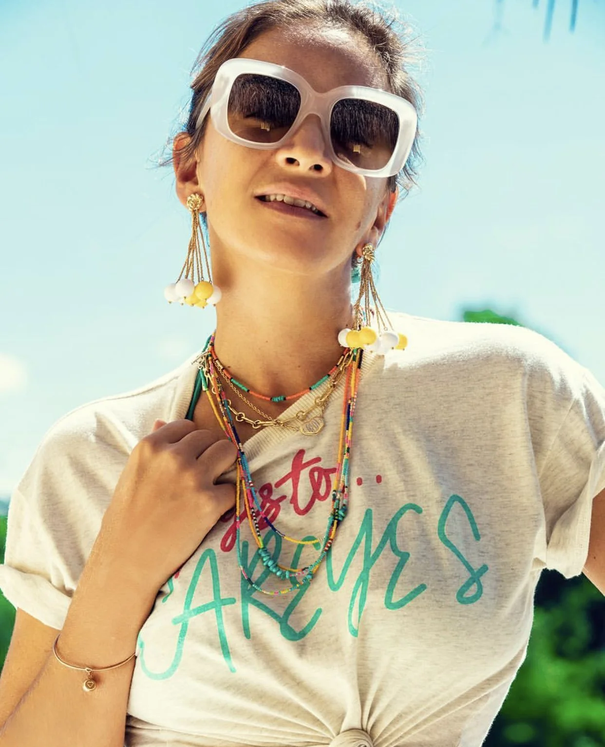Lauré Heriard Dubreuil wearing large white sunglasses, colorful Bunny Shapiro jewelry, and a casual LHD T-shirt, posing outdoors on a sunny day.