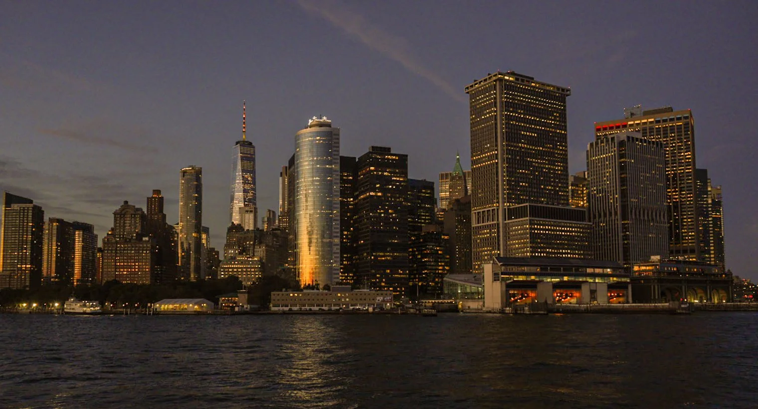 Nighttime view of New York City skyline with illuminated skyscrapers, including One World Trade Center, as seen from across the water.