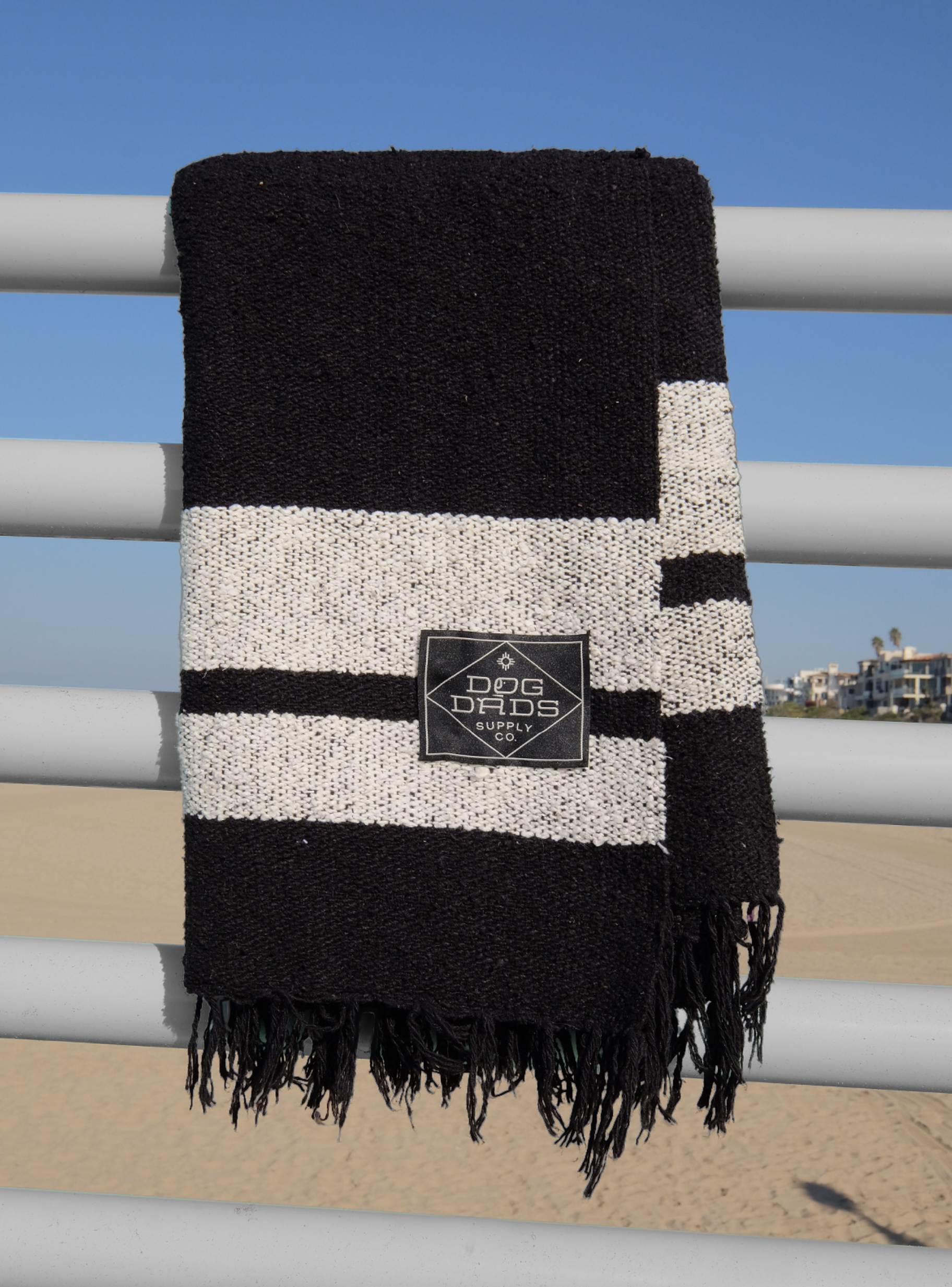 Black and white striped towel with a label that reads 'Dog Dads Supply Co.' hanging on a white railing at the beach.