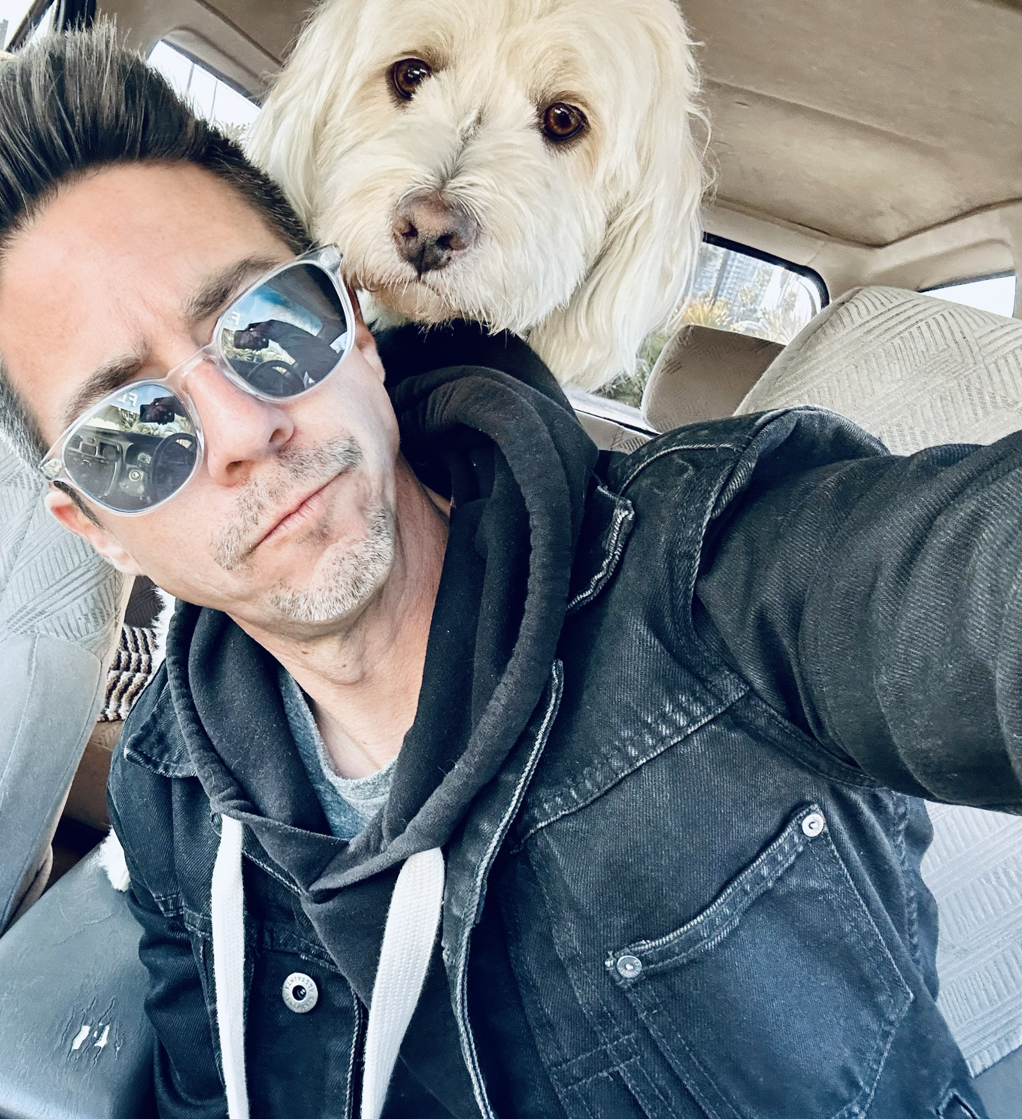 A man wearing sunglasses and a black leather jacket taking a selfie with a light-colored dog in the background, both inside a vehicle.