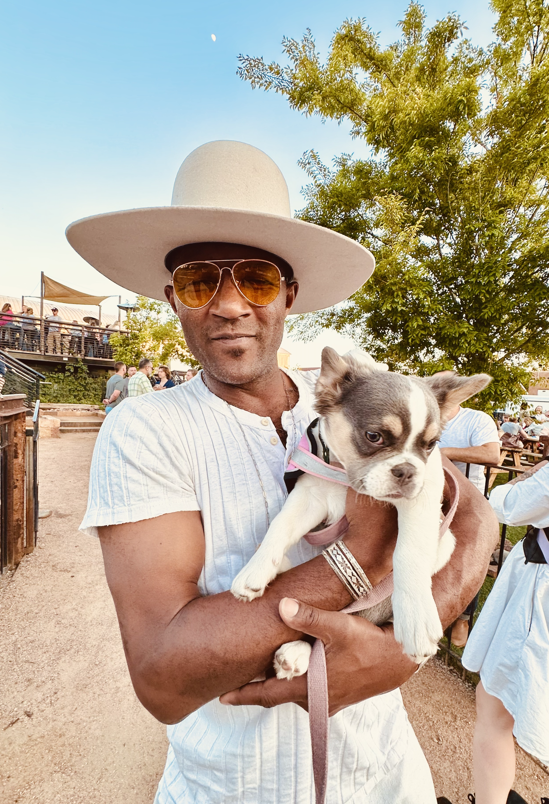 A man wearing a large wide-brimmed hat, sunglasses, and a white shirt holding a small puppy outside at a social gathering.