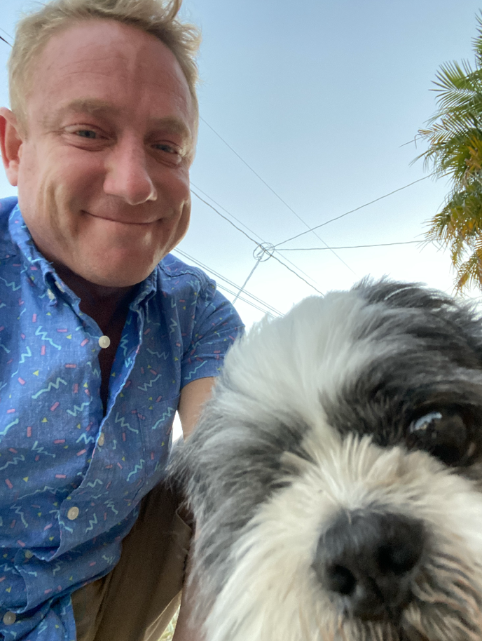 A smiling man with blond hair wearing a blue patterned shirt taking a selfie with a large black and white dog, outdoors with a clear sky, utility wires, and a palm tree in the background.