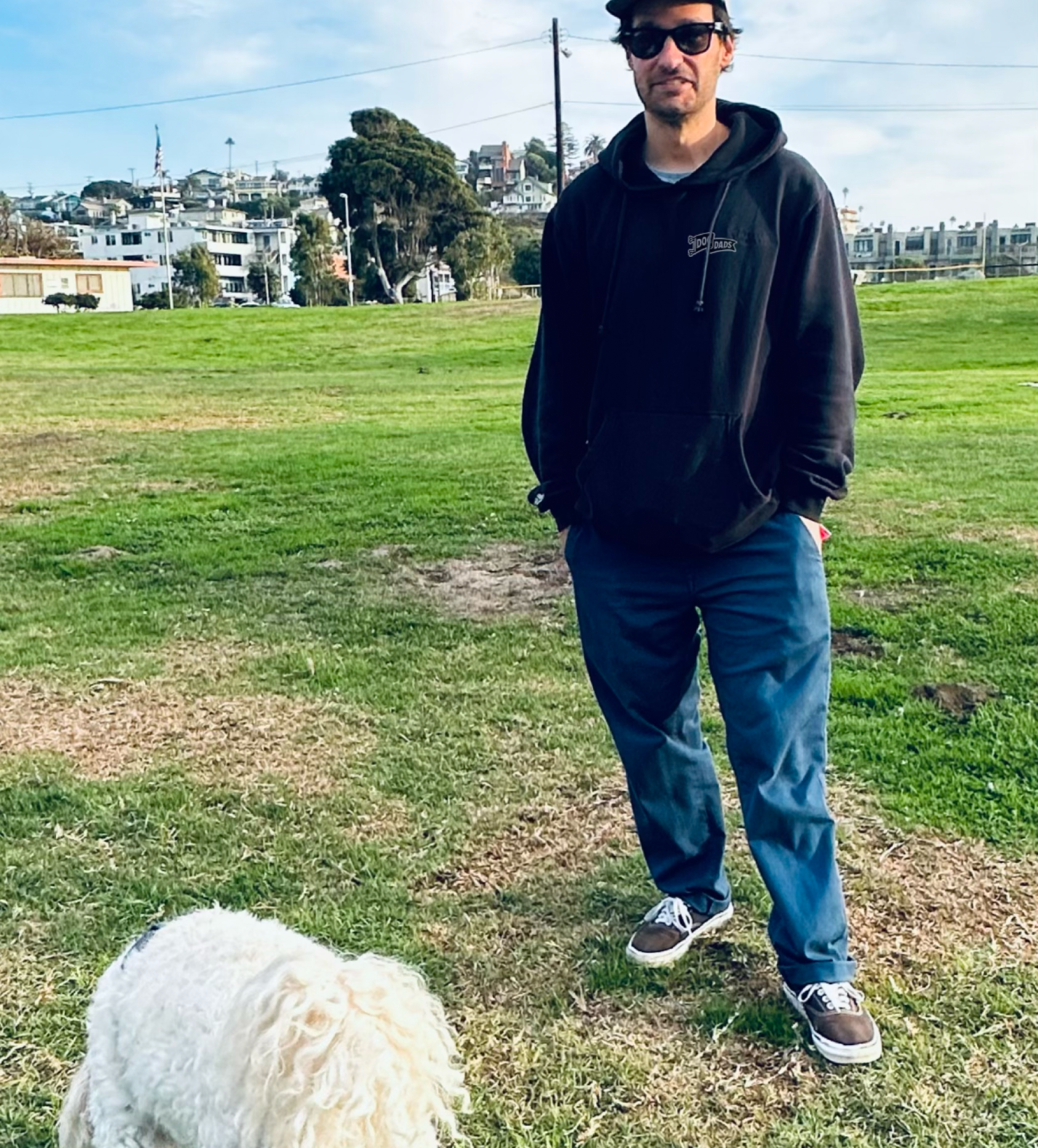 A man standing outdoors in a park with a white dog in the foreground and residential buildings in the background.