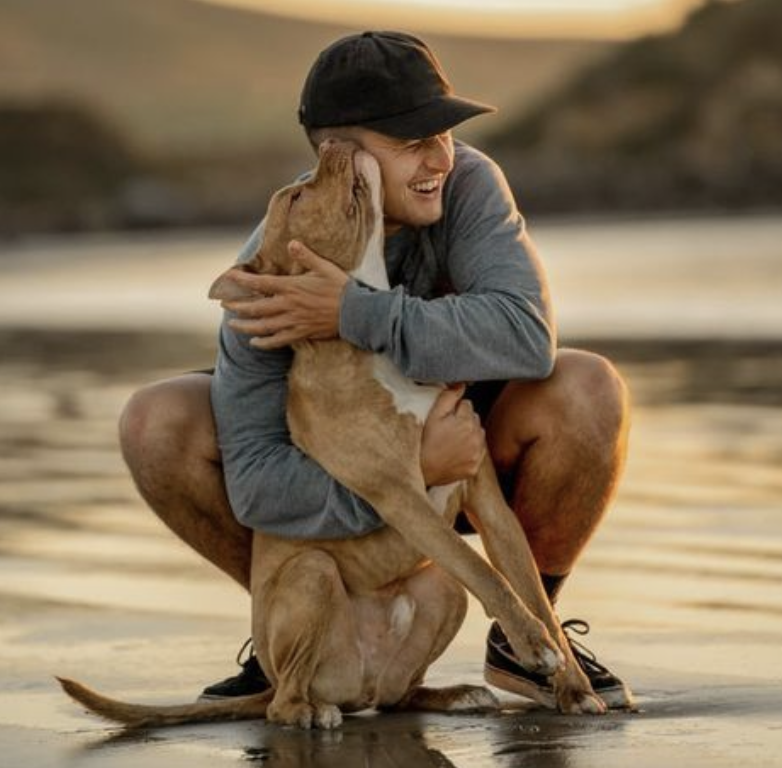 A person with a black cap and gray sweatshirt crouches on a beach, hugging a large brown and white dog, both smiling.
