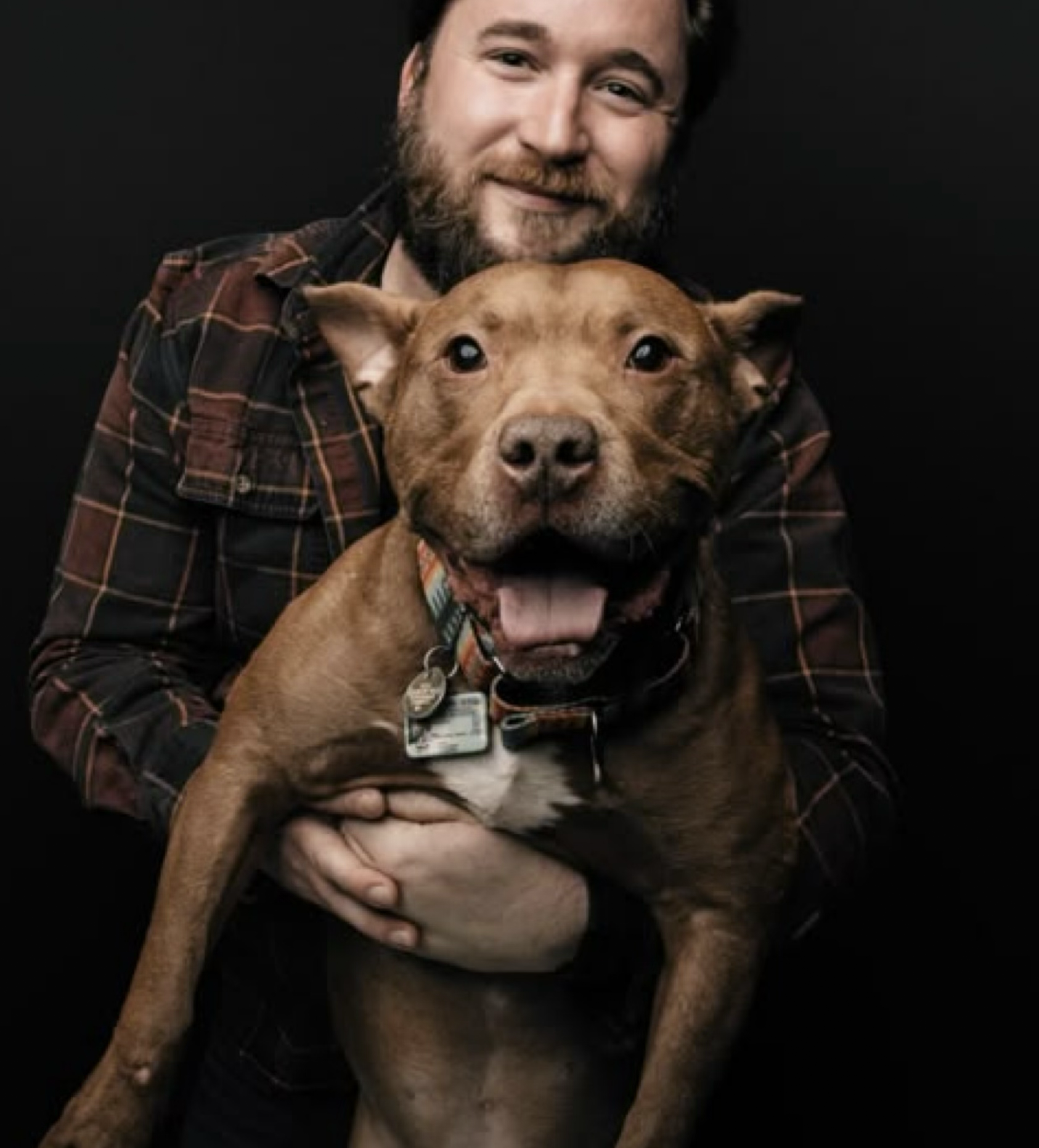 A man with a beard smiling while holding a happy brown dog with a colorful collar against a black background.