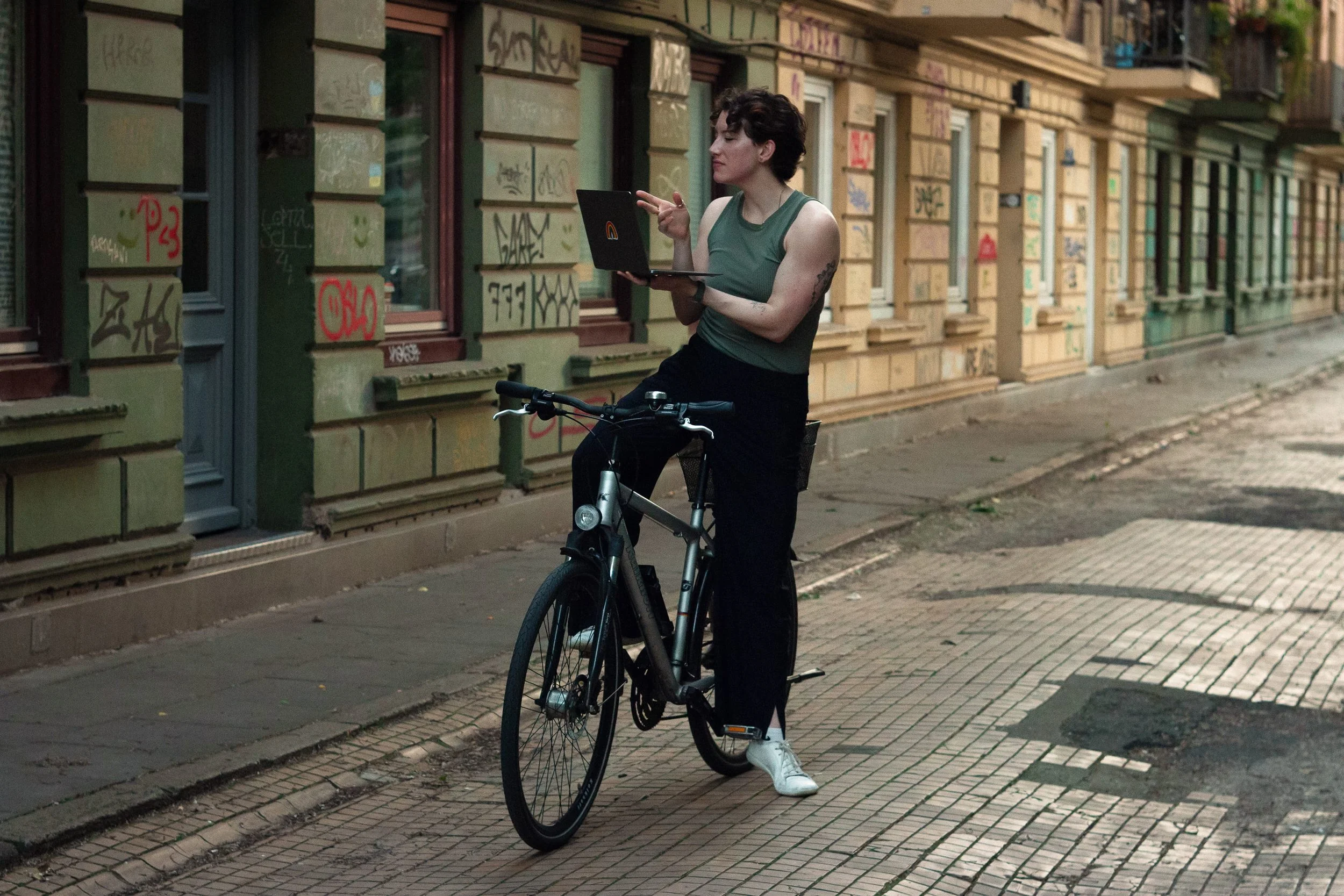 A woman with short dark hair and tattoos on her arm stands with a bicycle on a city sidewalk, using a laptop. She is wearing a green sleeveless top, black pants, and white sneakers. The background shows a building with graffiti on the walls.
