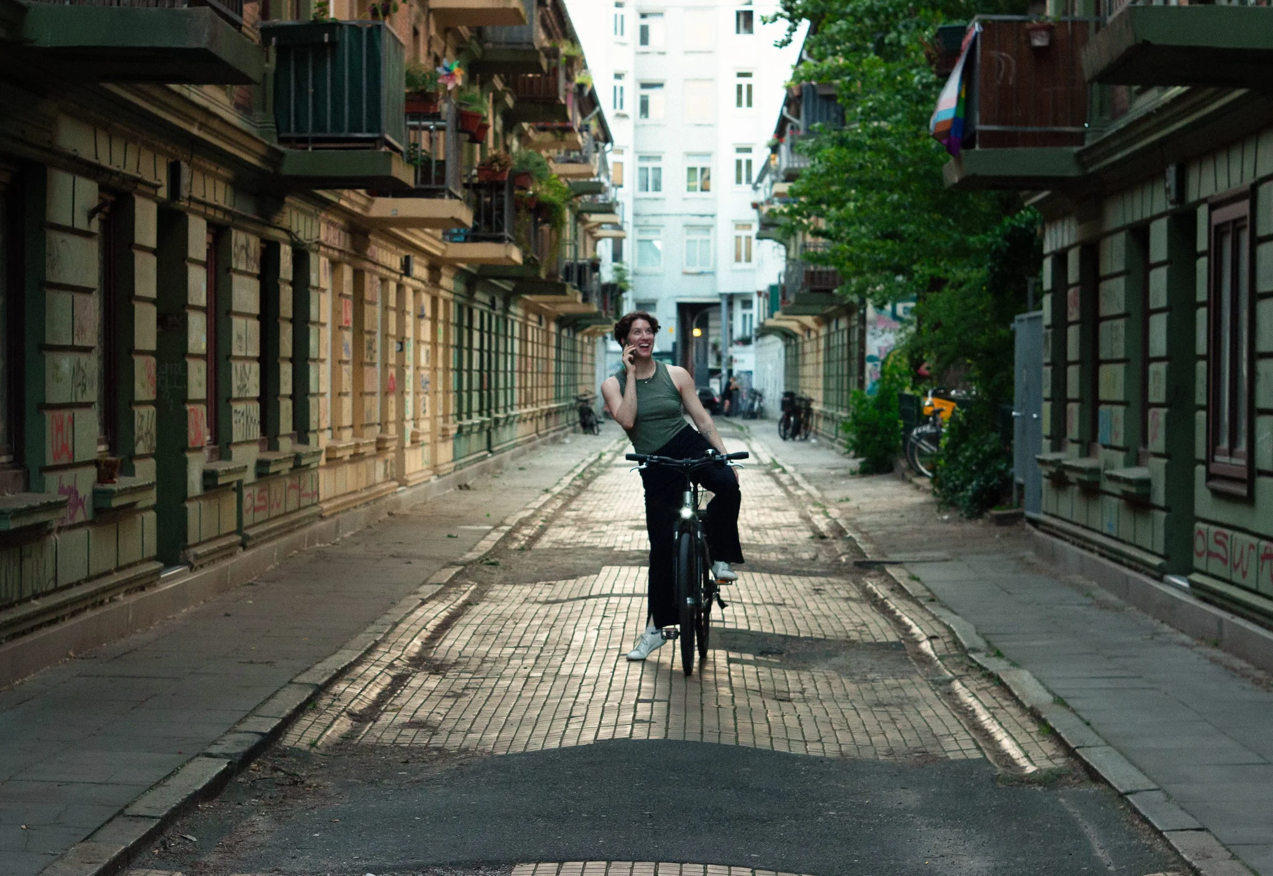 Woman riding a bicycle on a cobblestone alleyway, talking on her phone, smiling. Surrounding buildings are old with balconies and some graffiti, trees lining the street.