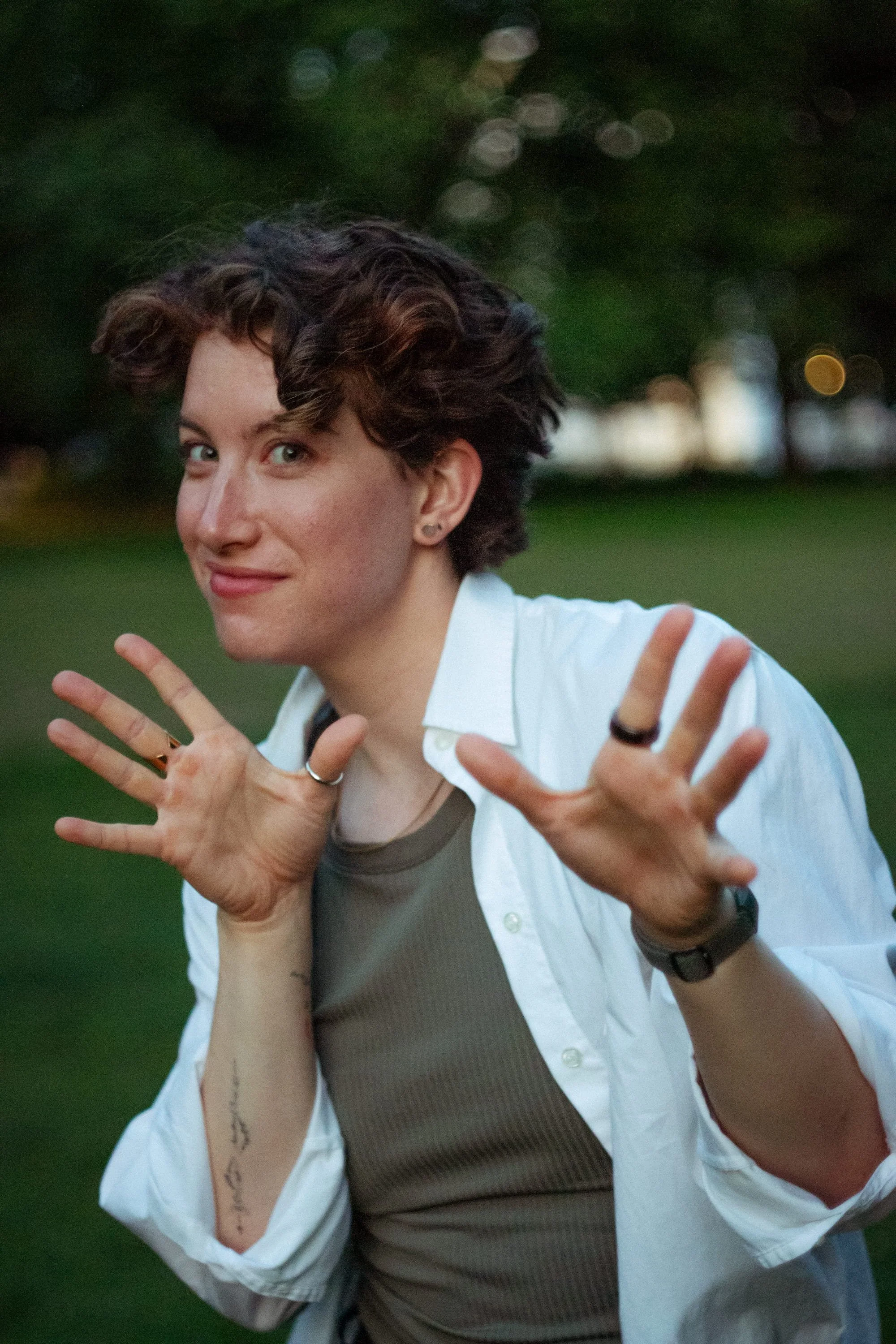 Person with short curly hair making a playful gesture outdoors in a park during evening.
