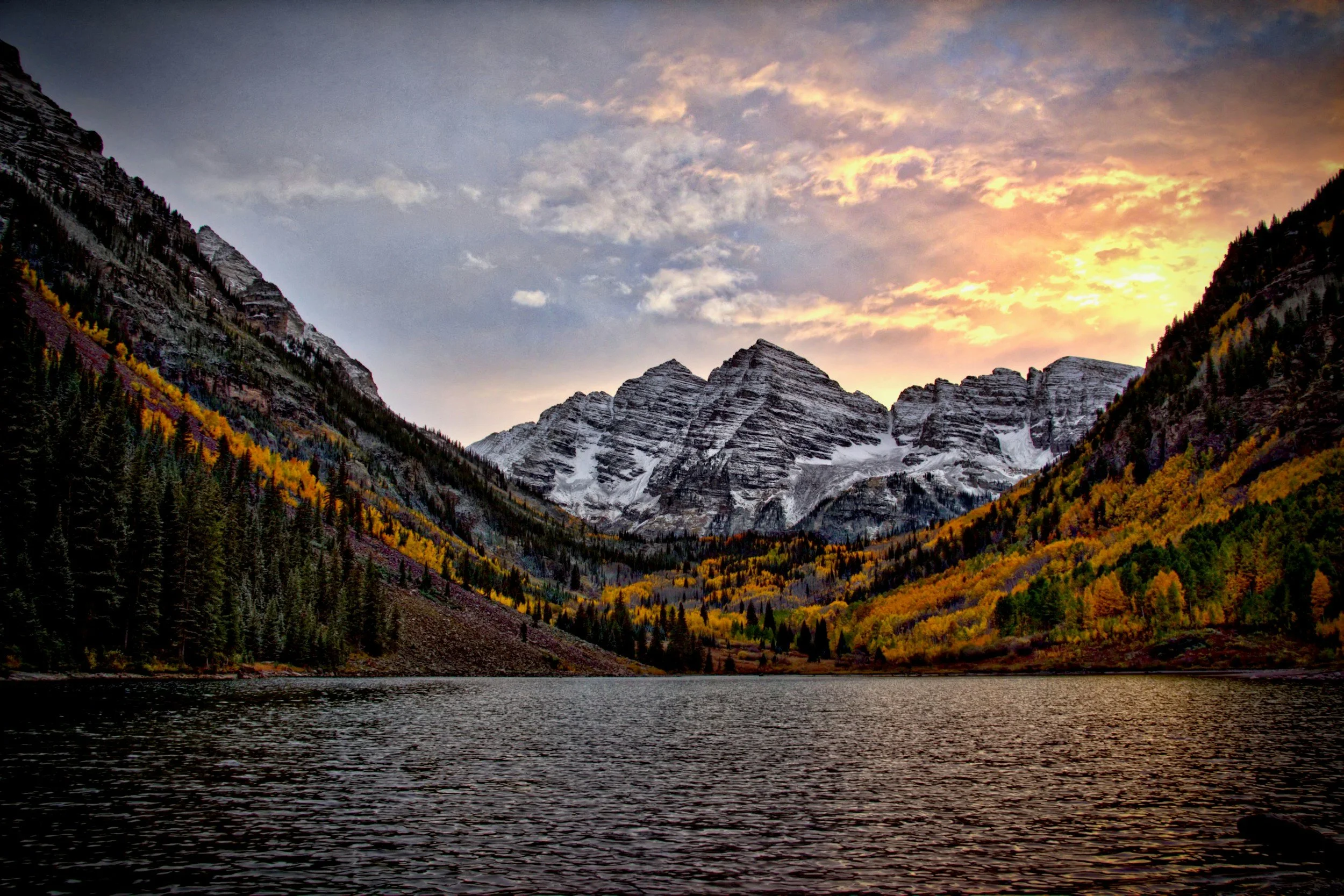 A scenic view of a mountain landscape during sunset, with snow-capped peaks, colorful autumn trees, and a body of water in the foreground.