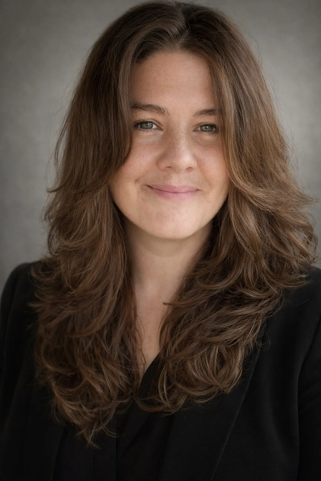 A woman with long, wavy brown hair, light skin, and blue eyes, wearing a black blazer, smiling softly at the camera against a neutral background.