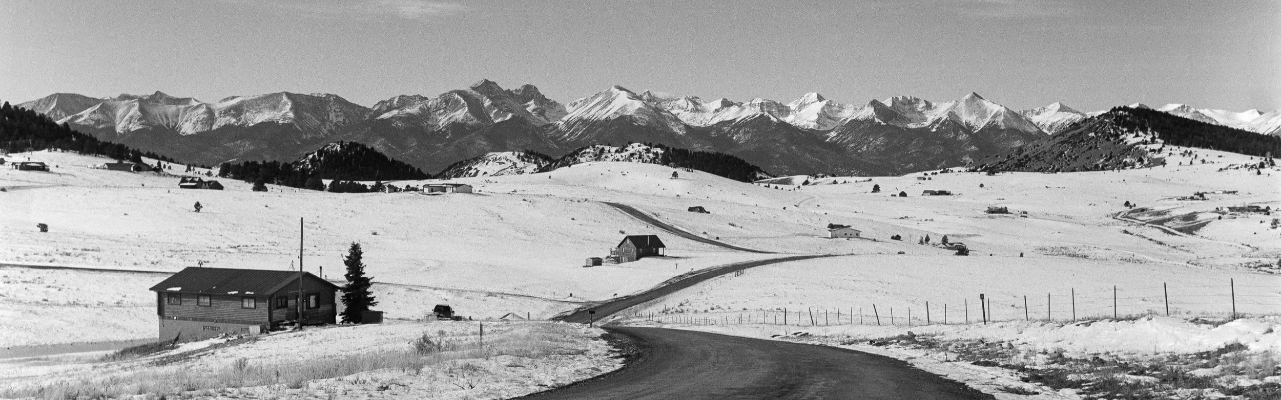 Scenic black and white landscape of snow-covered fields and rural houses with a mountain range in the background under a clear sky.