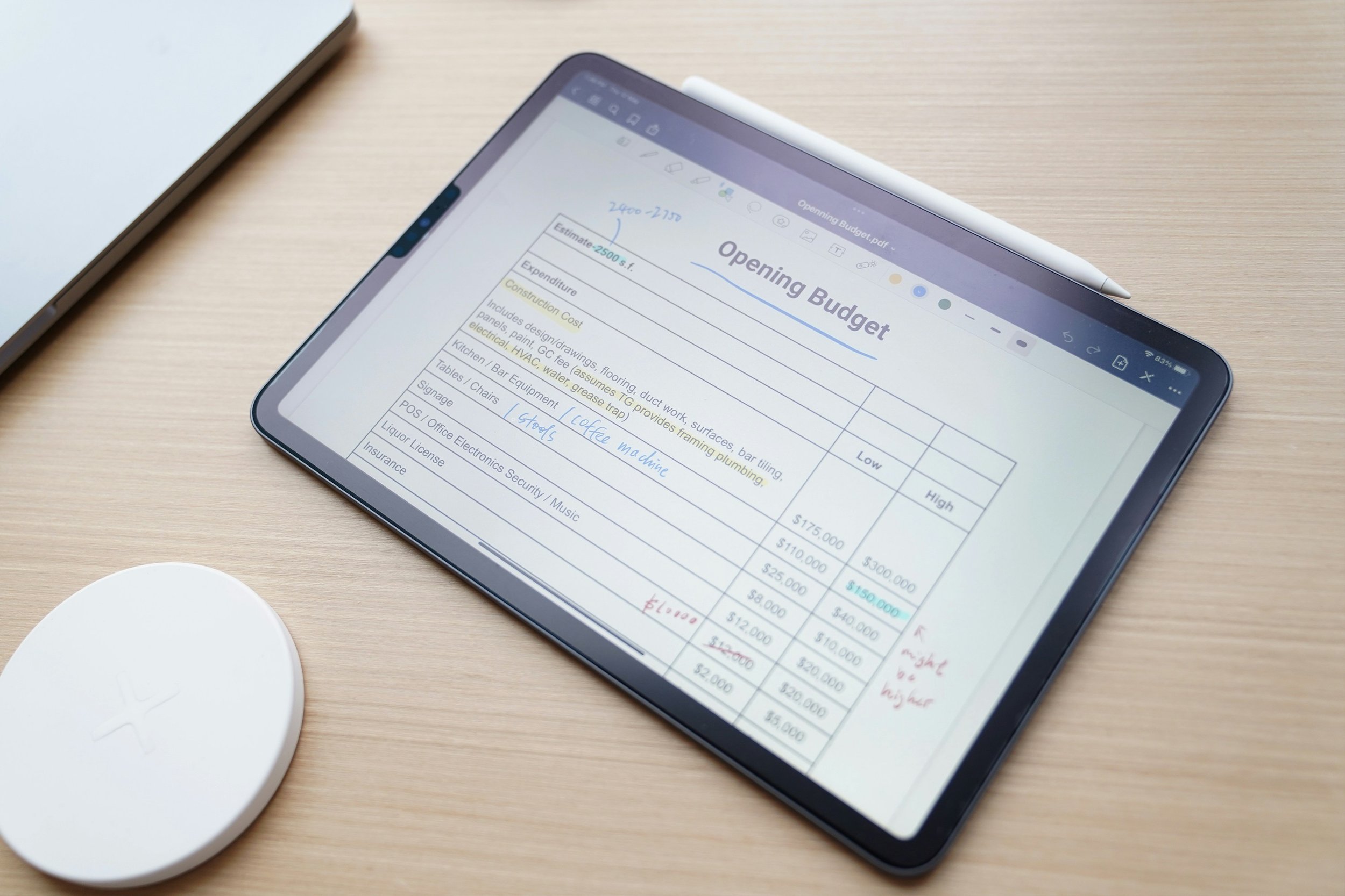 Tablet displaying an opening budget spreadsheet on a wooden desk with a white wireless charger nearby and a stylus placed above.